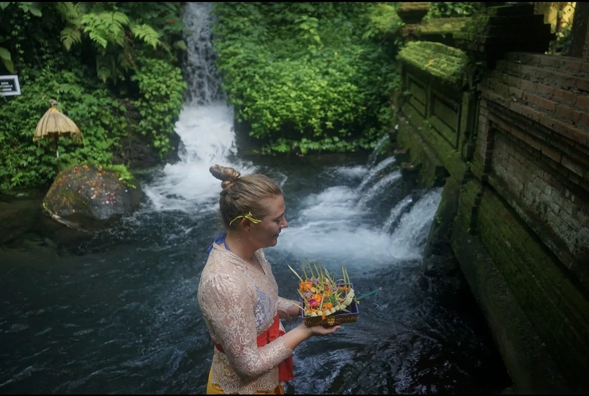 A woman in traditional Balinese attire holding a tray of colorful flowers, standing in front of a small waterfall in a lush, green setting.
