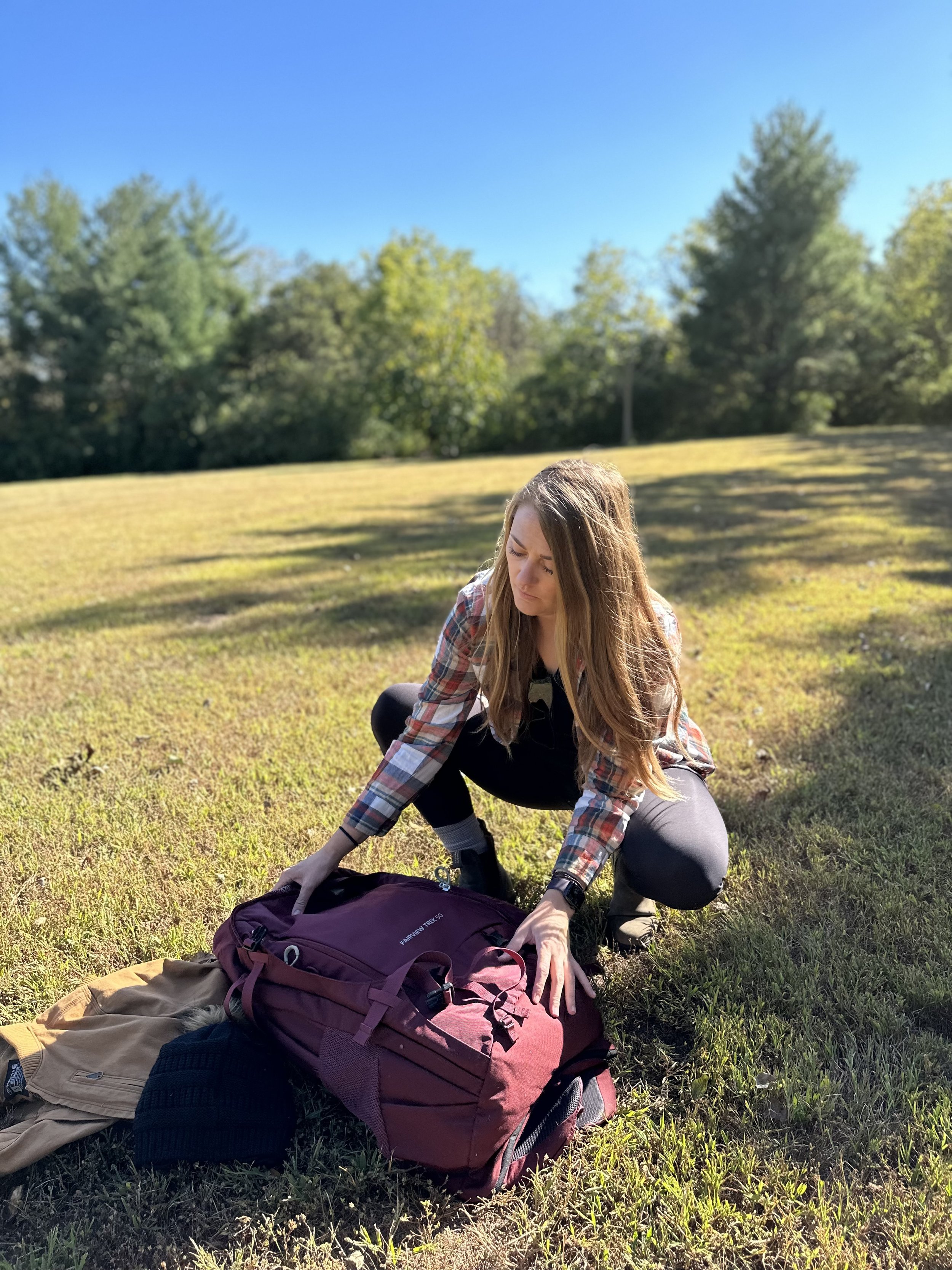 A woman with long brown hair wearing a plaid shirt and black pants is kneeling on green grass, packing a maroon backpack outdoors on a sunny day with a blue sky and trees in the background.