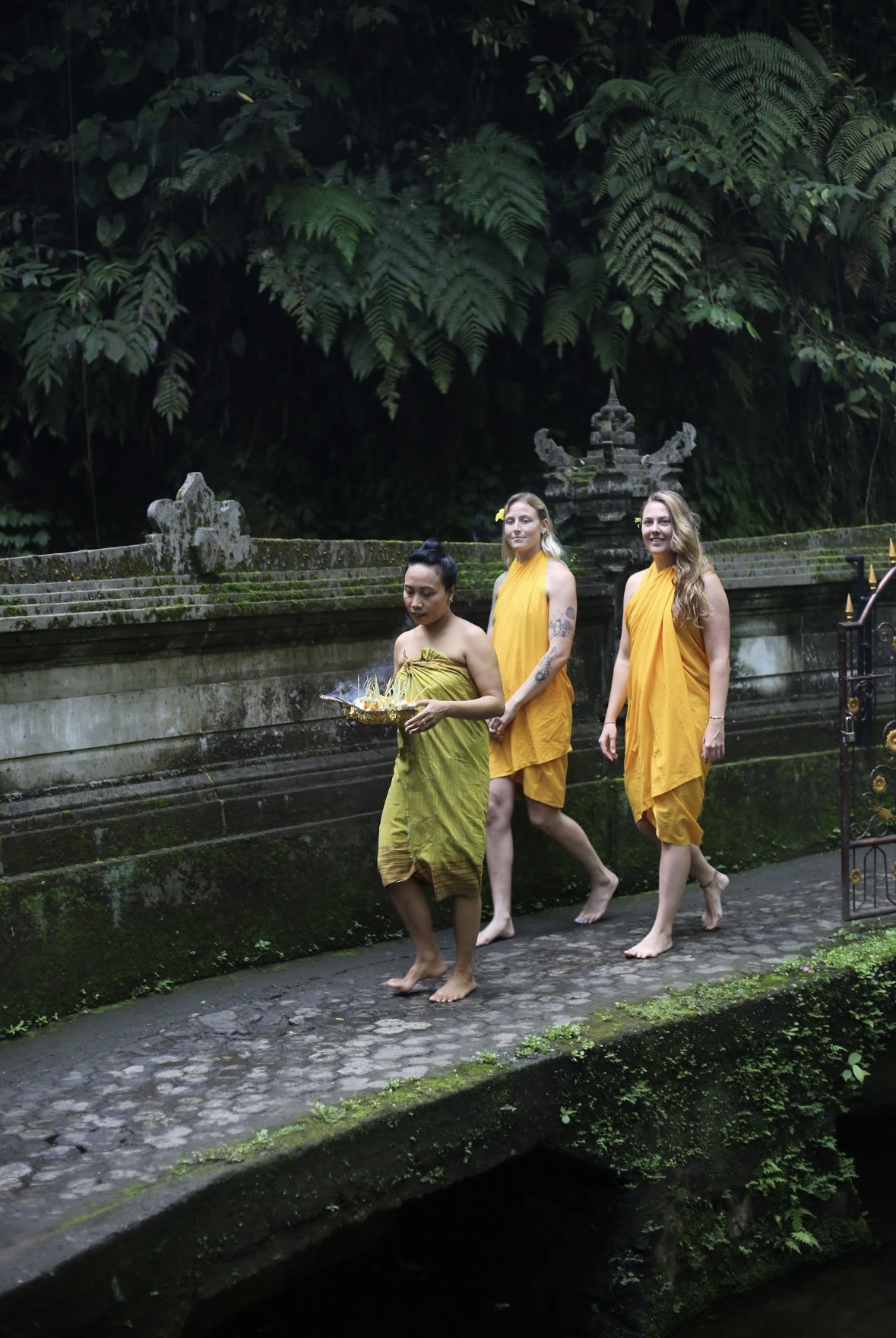 Three women in traditional orange and yellow robes walking barefoot on stone pathway in lush green tropical environment, with one woman holding a decorated tray, possibly for a religious or cultural ceremony.