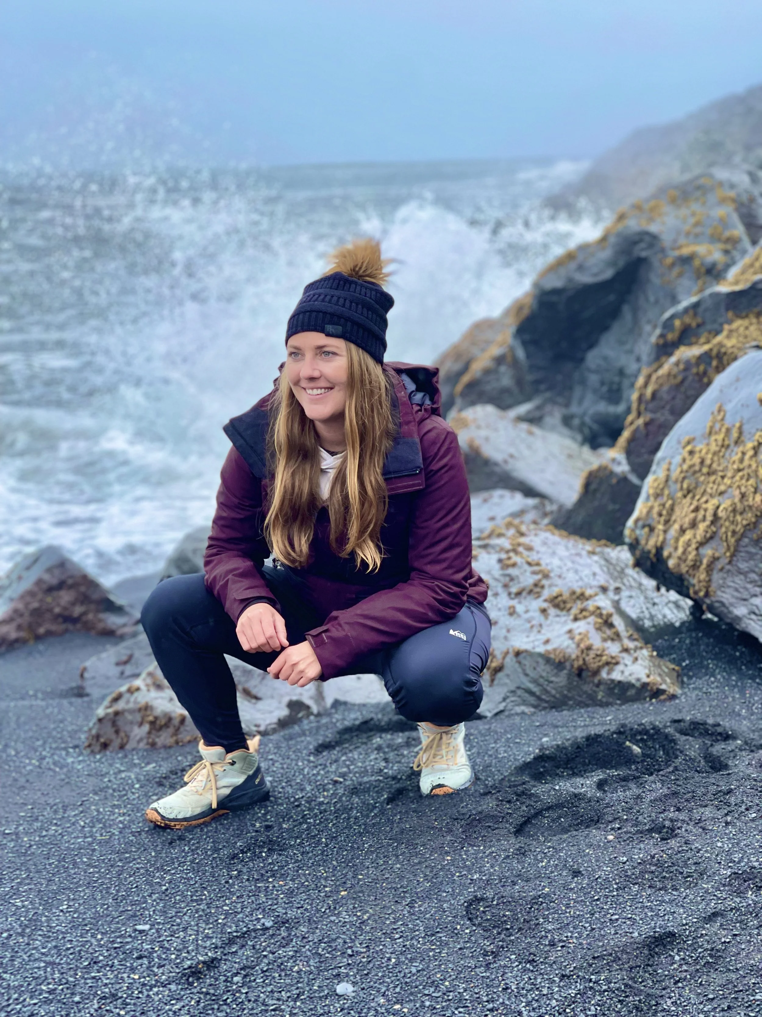 A woman crouching on a black sand beach near rocks, with ocean waves in the background, wearing a navy beanie, maroon jacket, and hiking shoes.