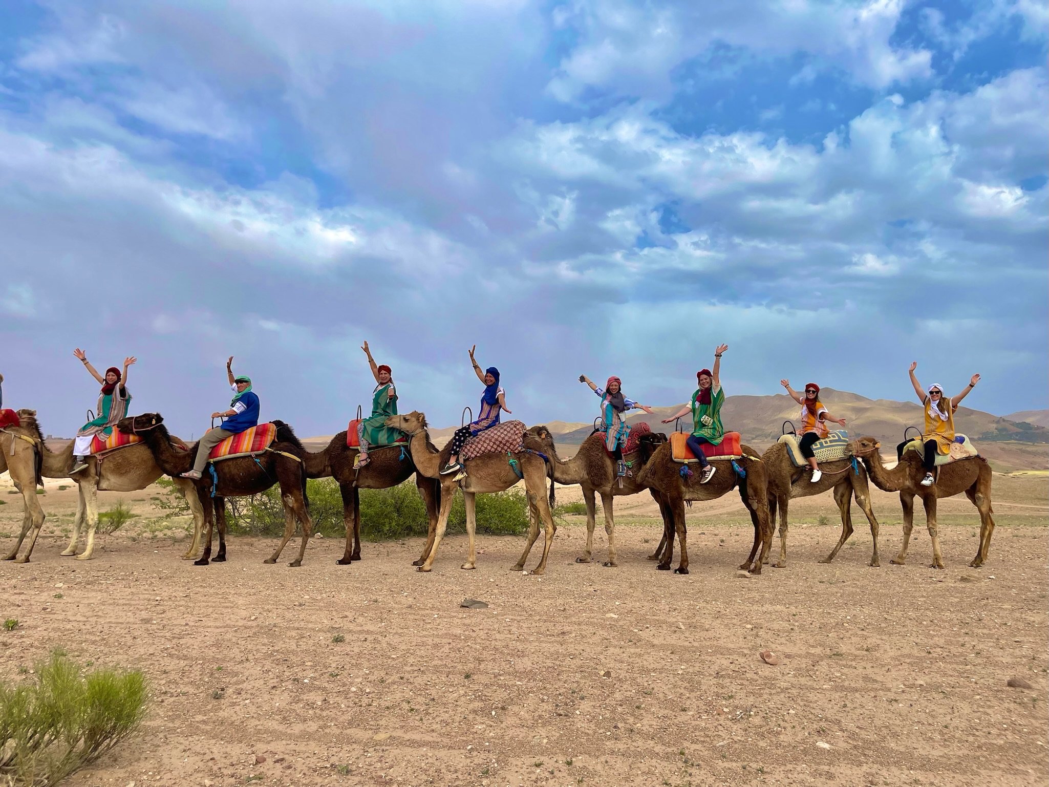 A group of people riding camels in a desert landscape with mountains in the background, under a partly cloudy sky.