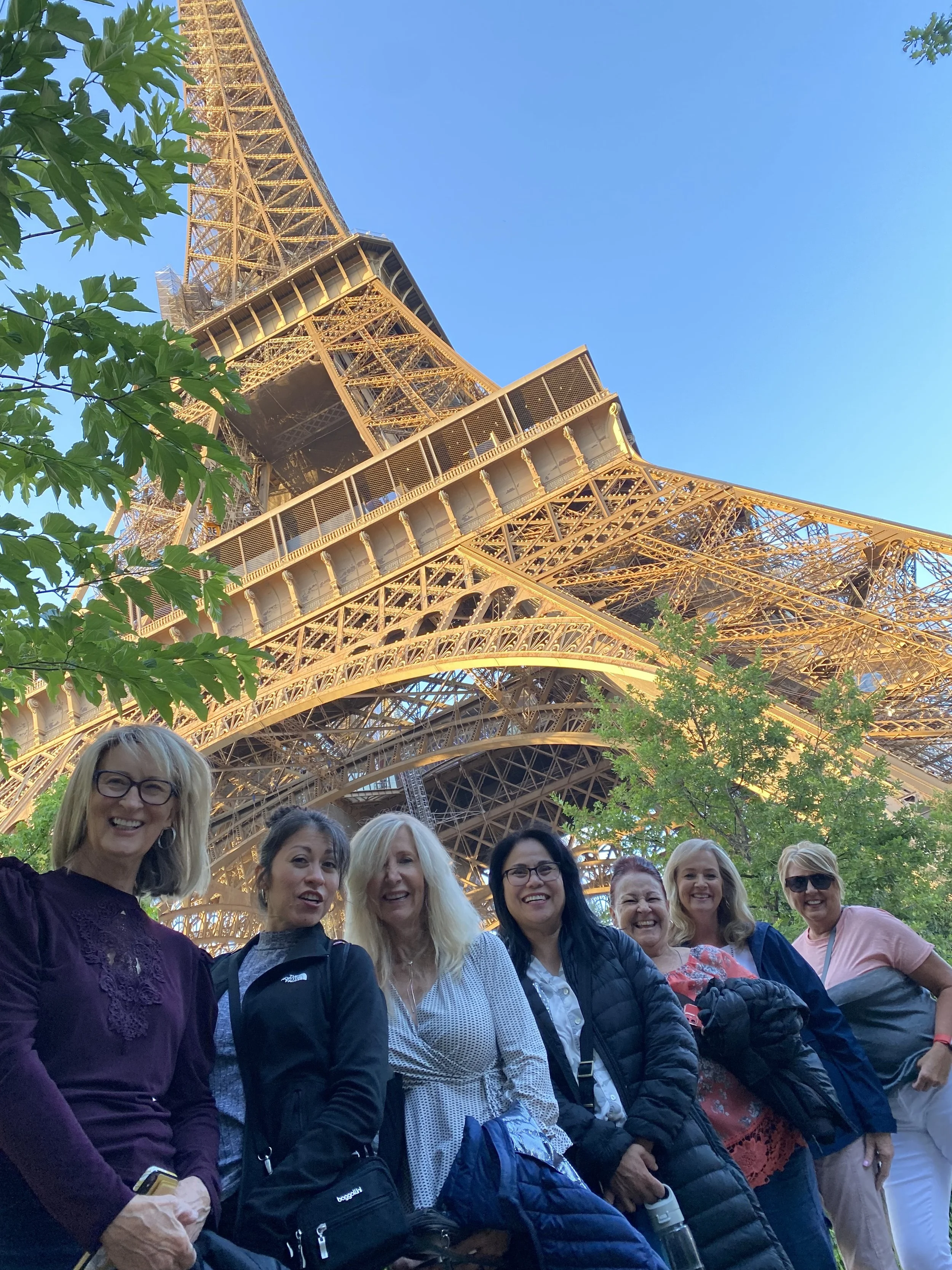 Group of seven women smiling in front of the Eiffel Tower in Paris, France, during daytime with clear blue sky and green trees.