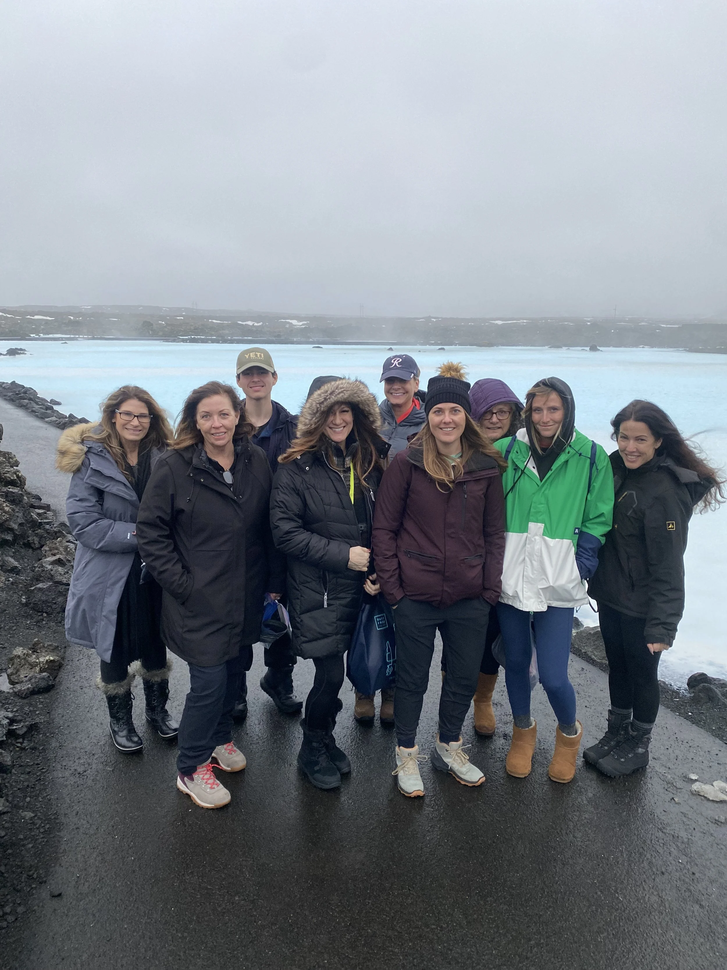 Group of nine people standing together outdoors near a body of water on a cloudy, overcast day, all wearing warm outdoor clothing.
