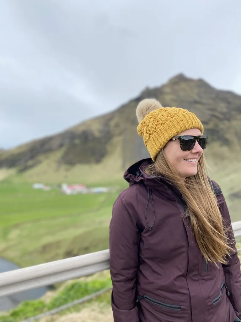 A woman in sunglasses and a mustard yellow knit beanie smiling outdoors with a mountain and green landscape in the background.