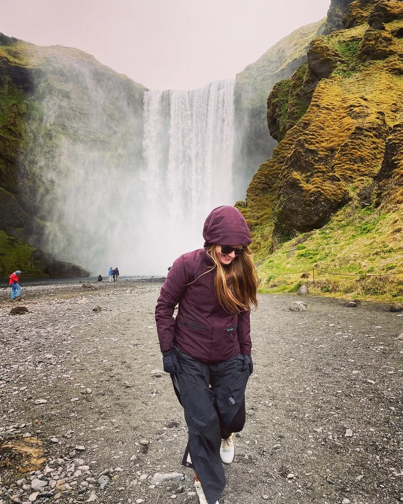 A woman in a purple jacket and black pants walking on a rocky trail near a waterfall, with moss-covered rocks on the right and a misty waterfall pouring down from a high cliff in the background.