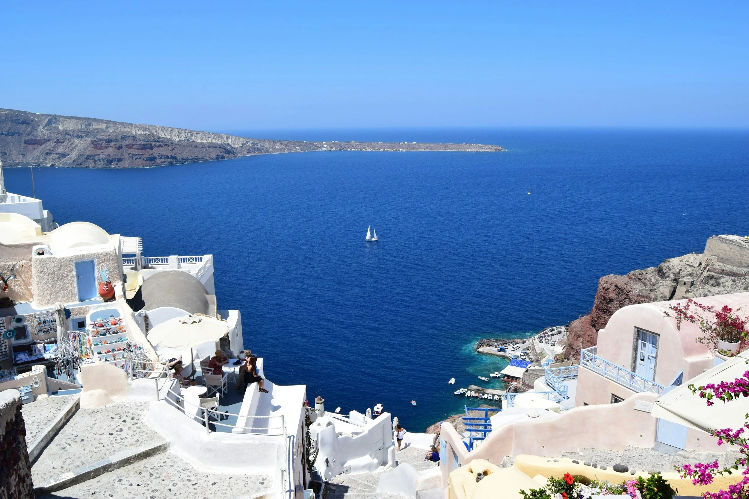 A scenic view of Santorini, Greece with white buildings on cliffs overlooking the deep blue Aegean Sea. There are boats sailing in the water and a clear sky above.