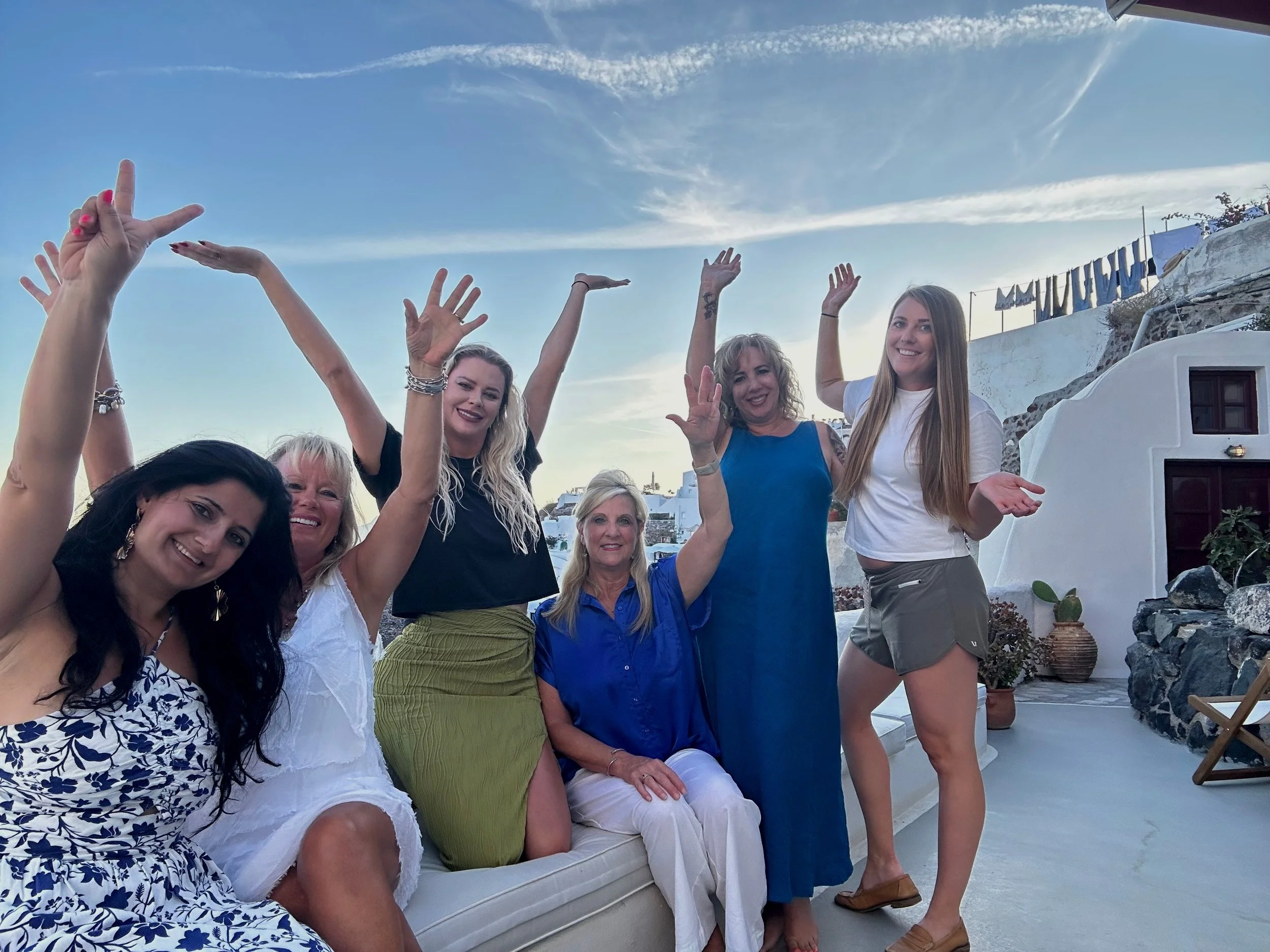 Group of six women celebrating outdoors on a terrace with white walls and a blue sky.