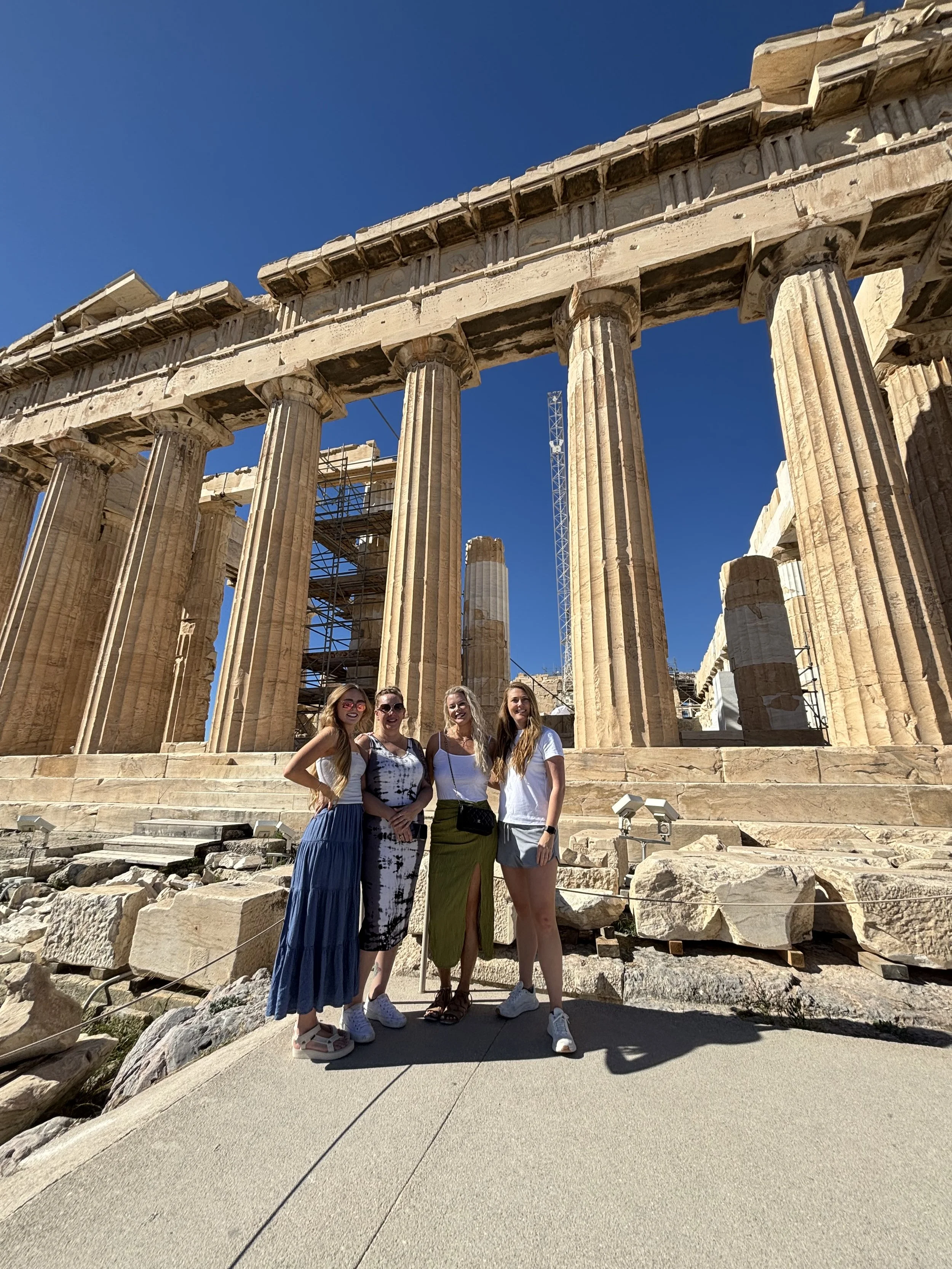 Four women standing in front of ancient Greek ruins of the Parthenon, with scaffolding and a crane in the background on a sunny day.