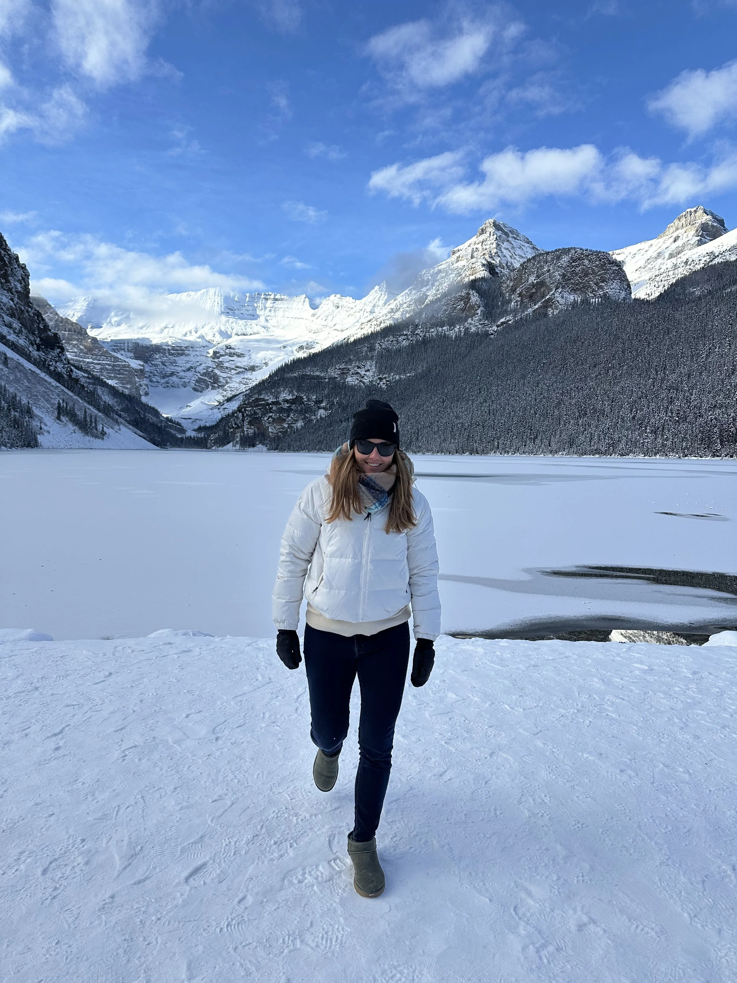 Woman wearing winter clothing running on snow near a frozen lake with snow-capped mountains in the background under a blue sky