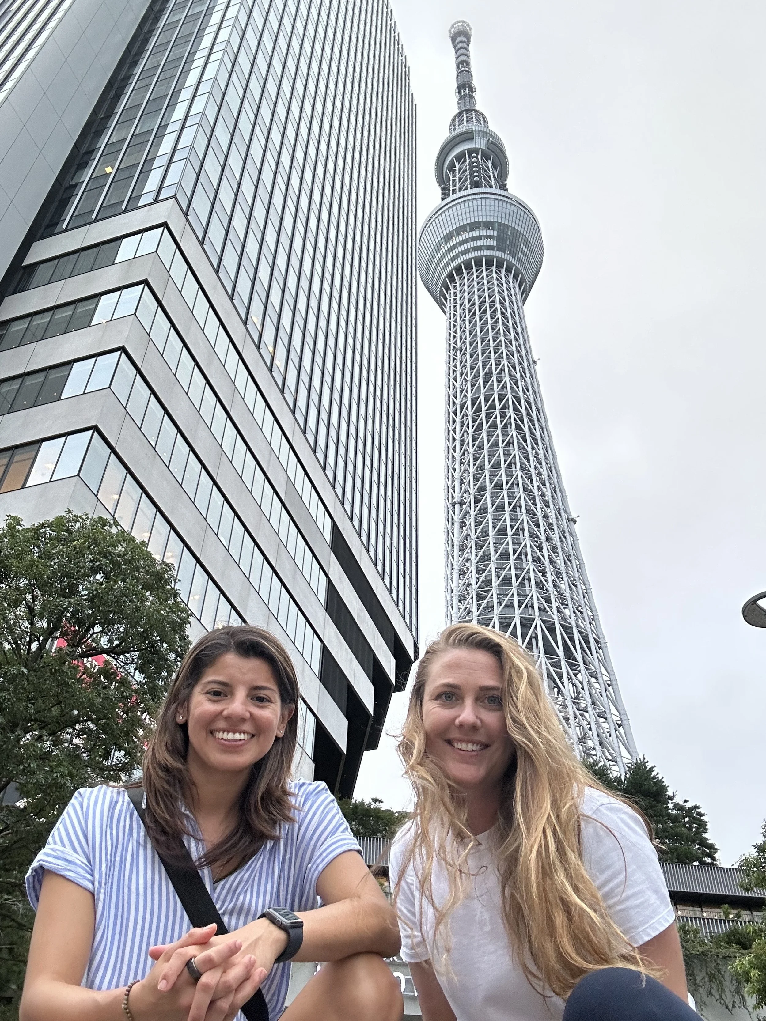 Two women smiling in front of the Tokyo Skytree tower with a modern building behind them.