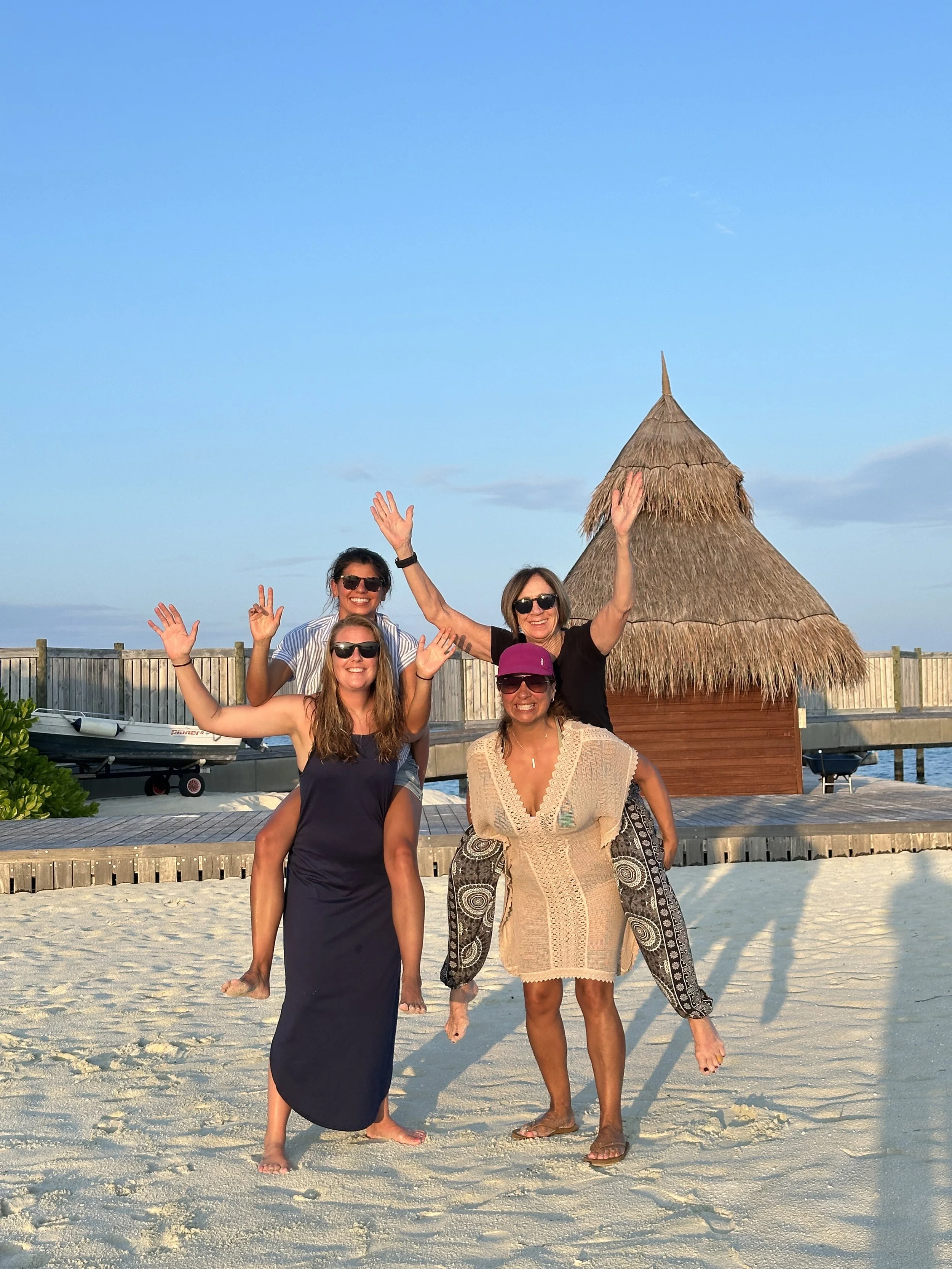 Four women smiling and waving on a sandy beach with a thatched-roof hut and a clear blue sky in the background.