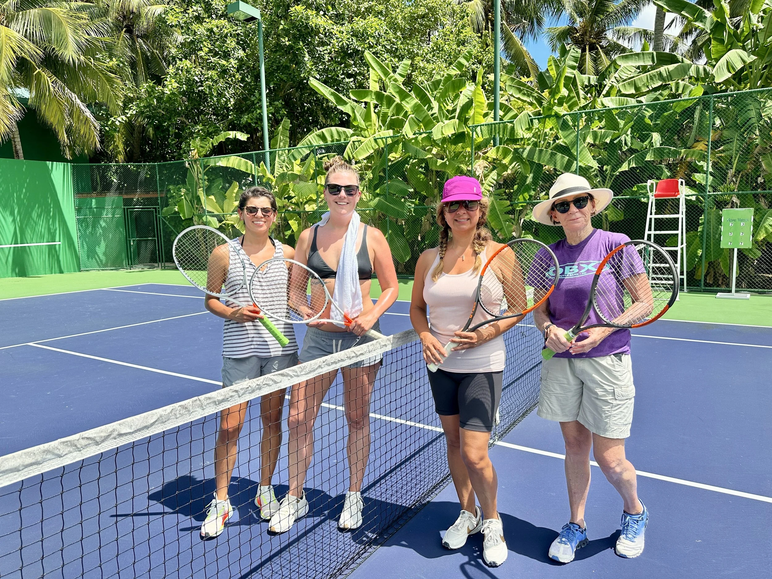 Four women standing on a tennis court holding tennis racquets, with a net in front of them, surrounded by lush green trees and banana plants.