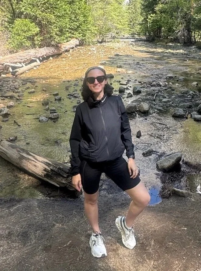 A woman wearing black sunglasses, a black jacket, black shorts, and running shoes standing on a dirt path near a rocky creek in a wooded area.
