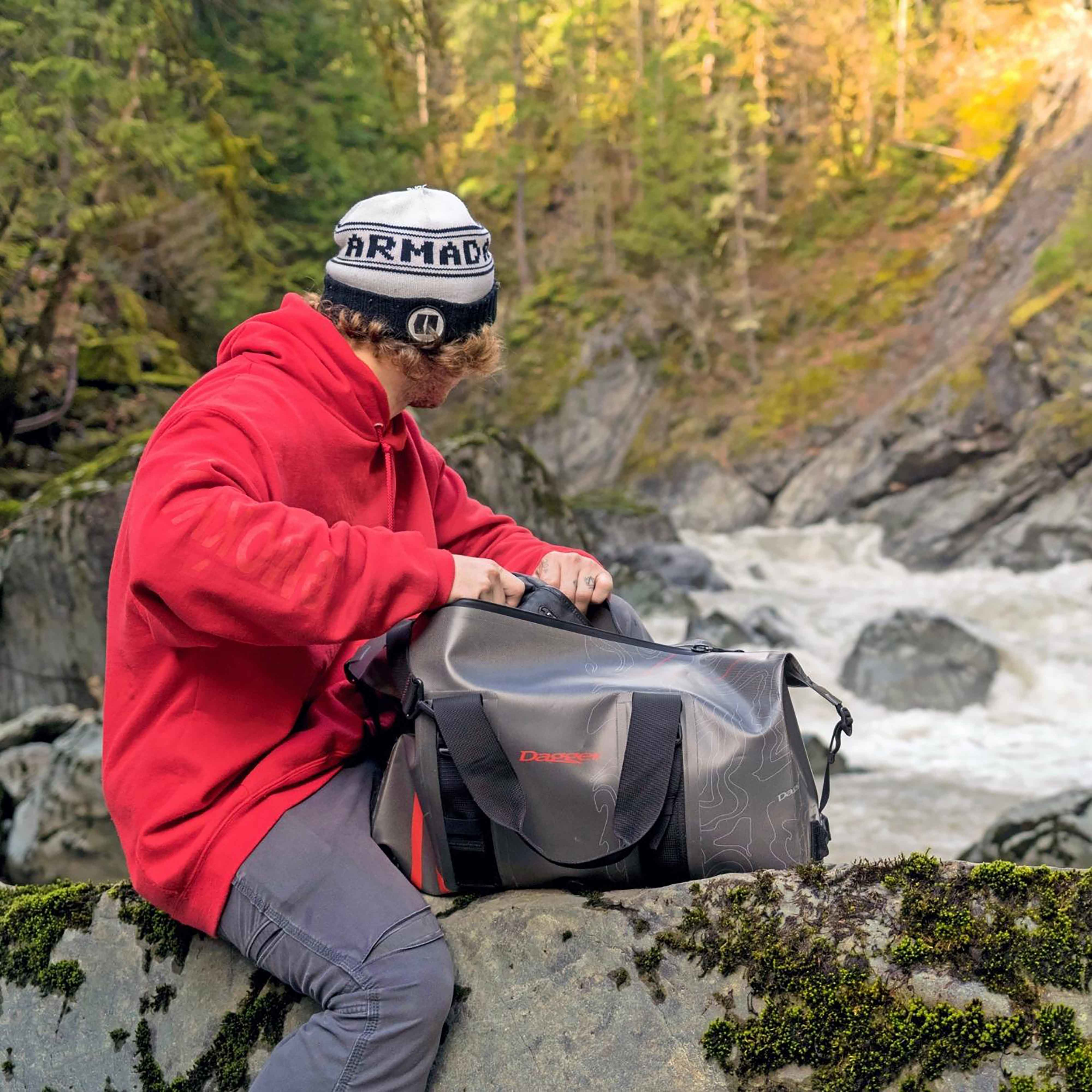 Image of man sitting in the wilderness next to a flowing creek opening his On-tap duffel dry bag from Dagger.