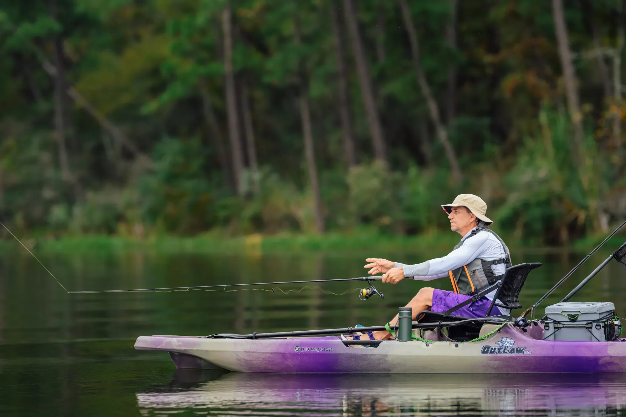 A man fishing from a purple kayak on a calm lake with a forested background.