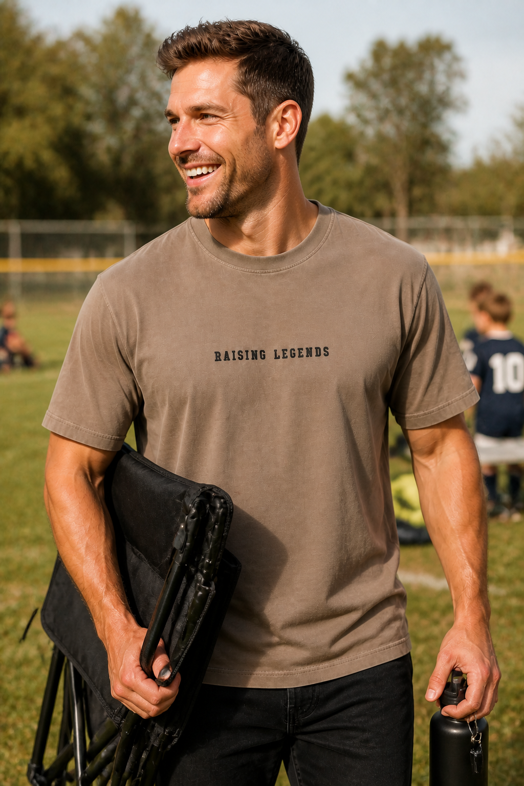 A smiling man in a beige t-shirt that says 'Raising Legends,' holding a folded portable chair and a water bottle, on a grassy field with children sitting on a bench in the background.