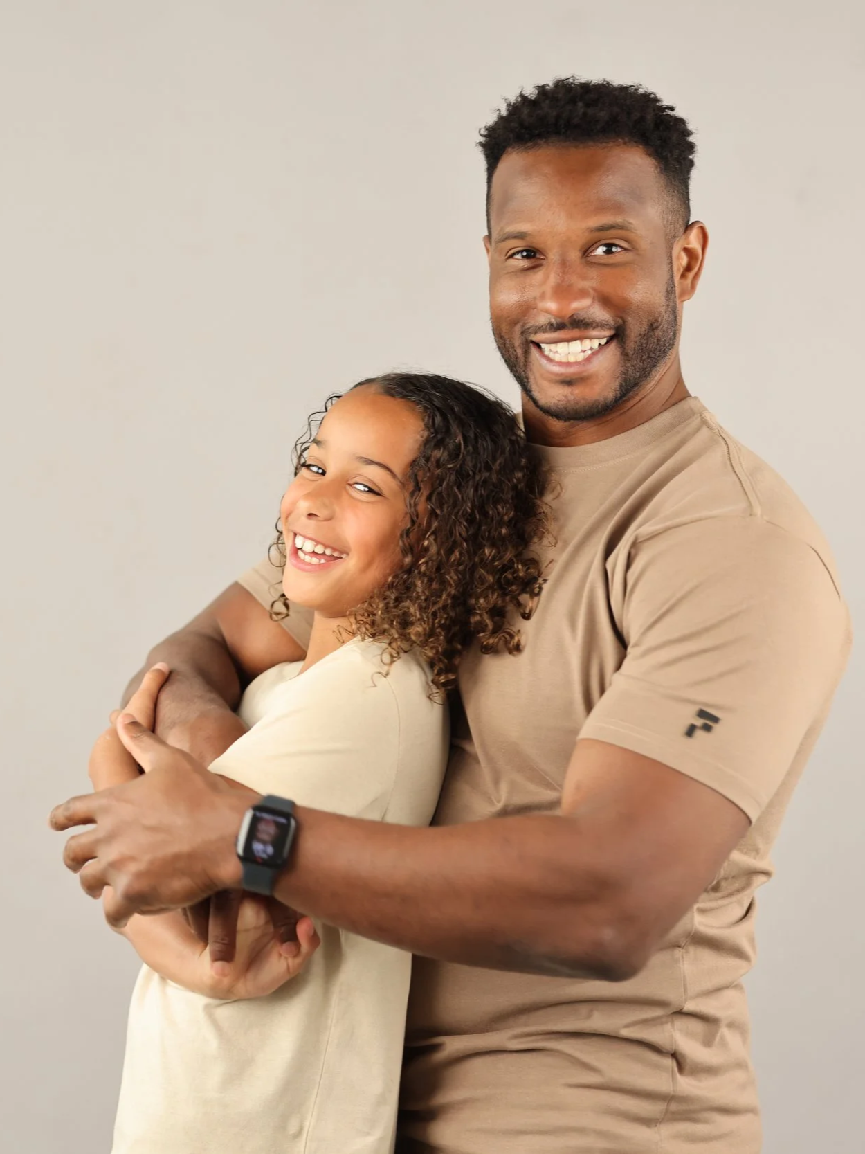 A man and girl smiling and hugging each other against a plain background. The man is wearing a beige t-shirt and a smartwatch, and the girl has curly hair and is wearing a light-colored shirt.