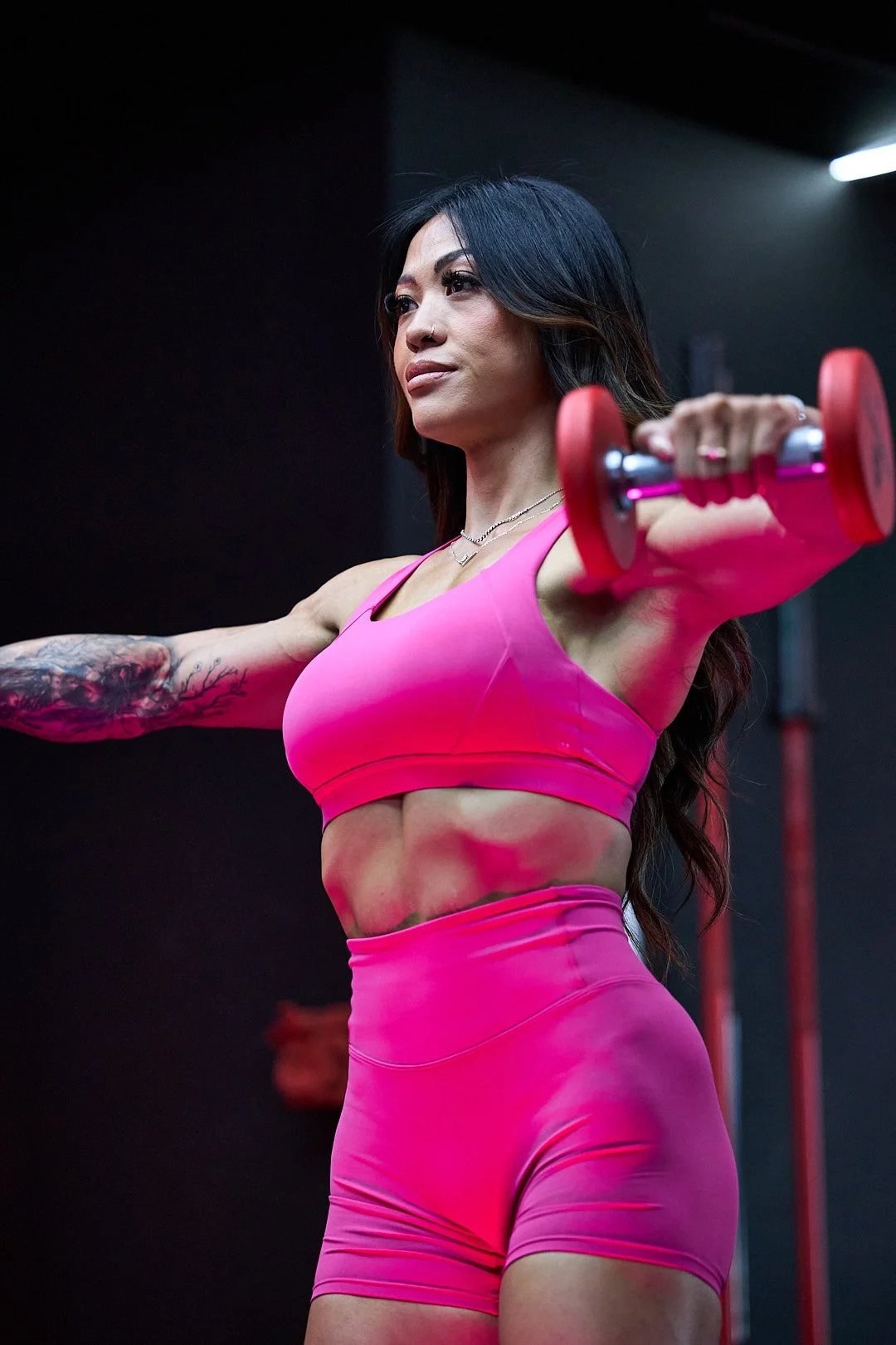 A woman in a pink sports bra and shorts holds a small pink dumbbell in a gym.