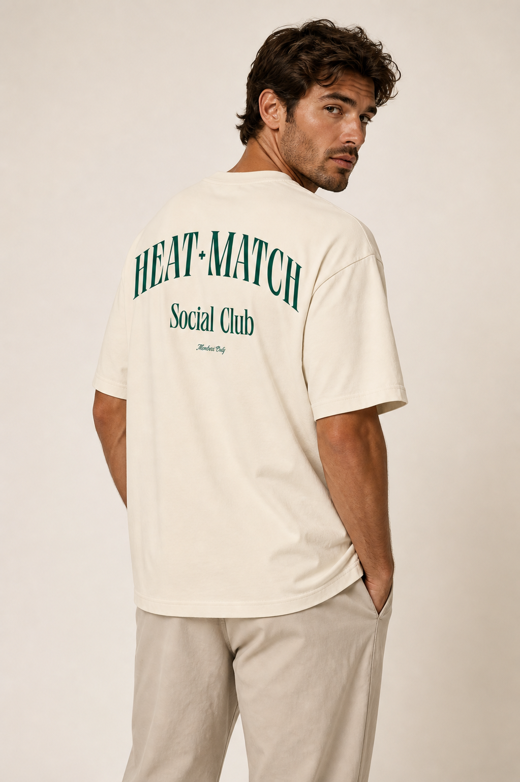 A man with dark hair wearing a beige T-shirt and matching beige pants. The back of the T-shirt has green text that reads "HEAT + MATCH," "Social Club," and a smaller cursive line that says "Member Only."