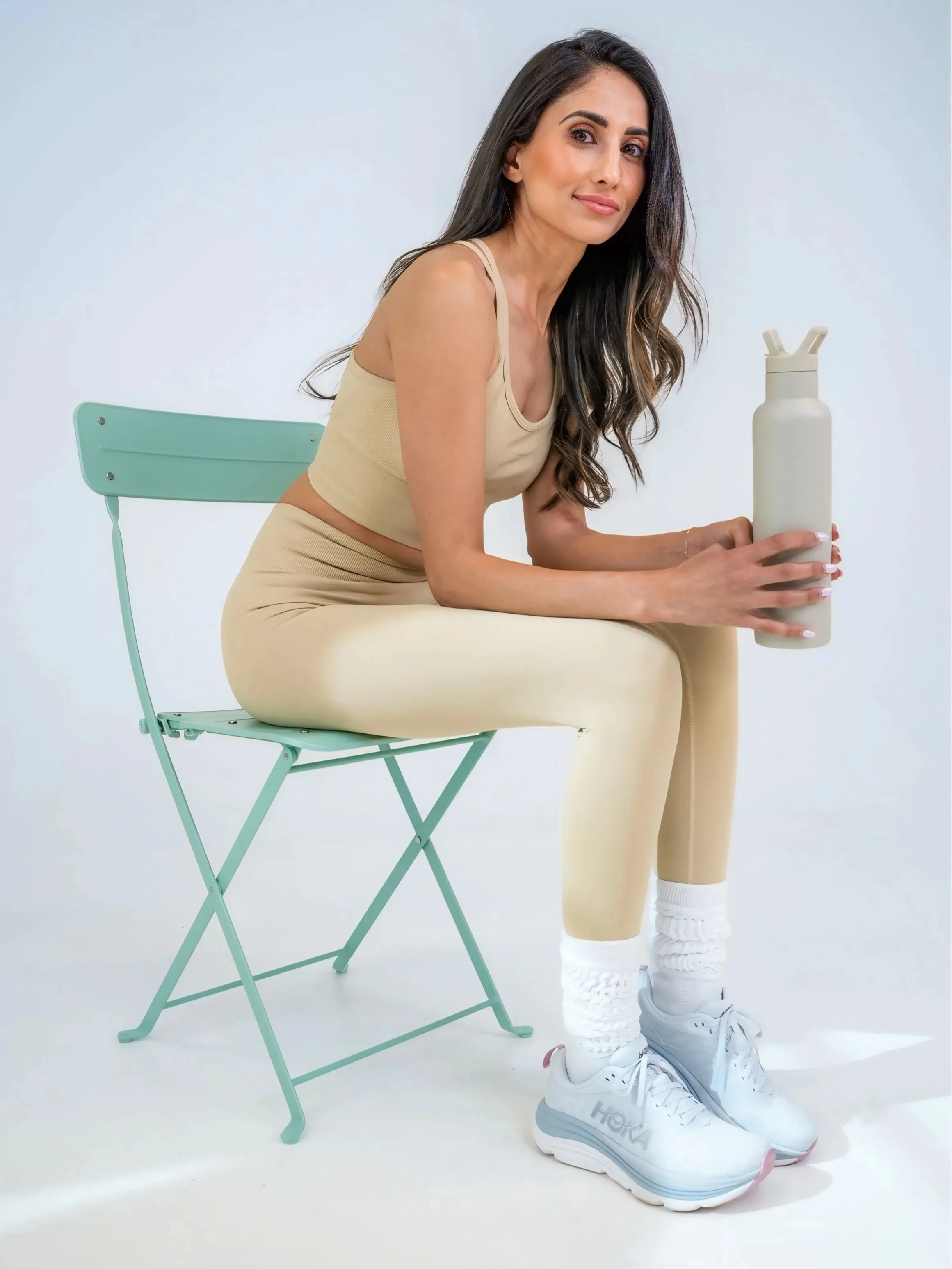 A woman with long dark hair wearing beige athletic wear and white sneakers, sitting on a light green metal folding chair, holding a beige water bottle, against a plain white background.