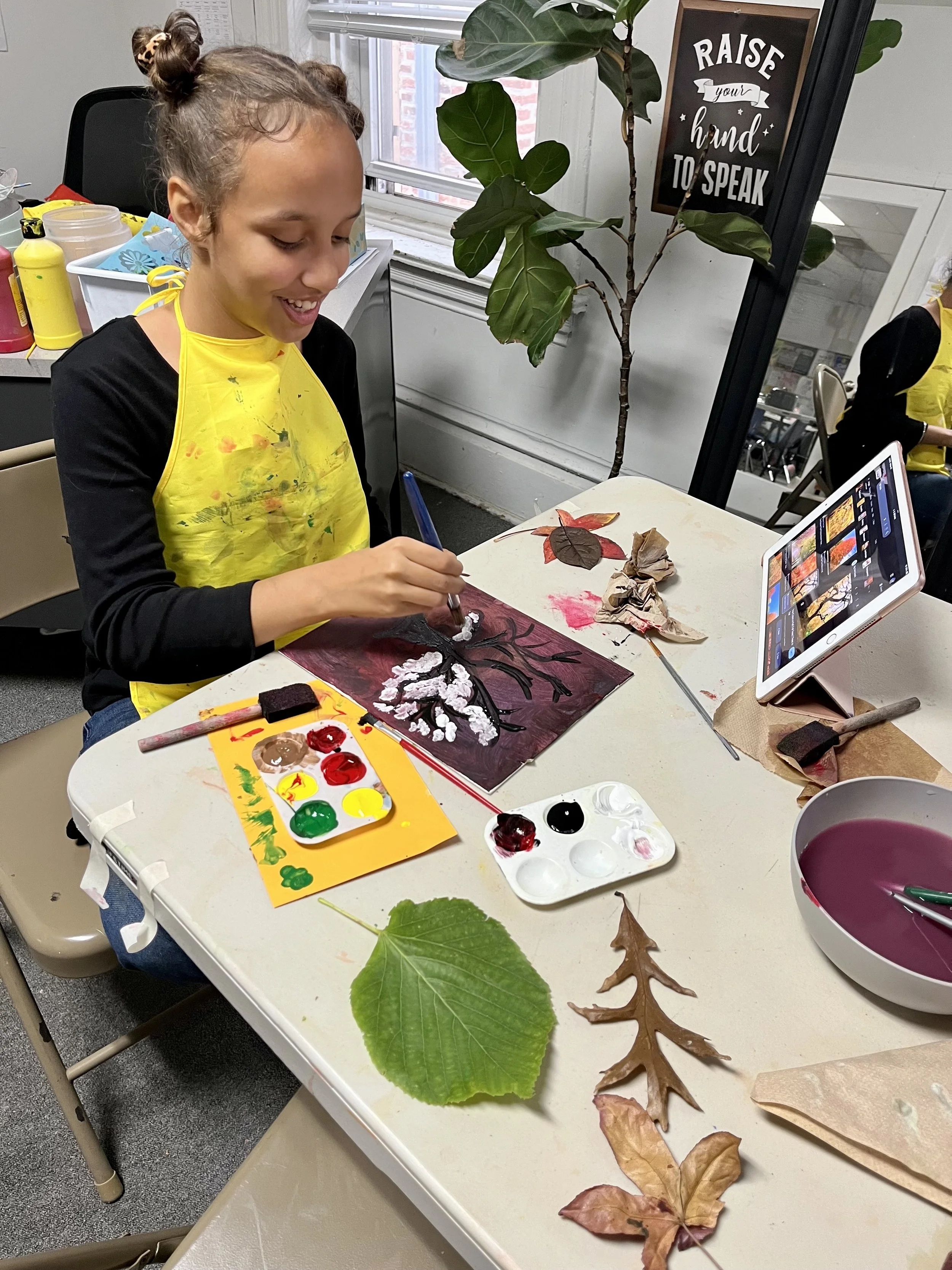 Young girl seated at a table painting a leaf with acrylic paints. The table has various leaves, watercolor paints, a bowl of water, paper towels, and a tablet showing a tutorial. A sign on the wall reads 'Raise your hand to speak.'