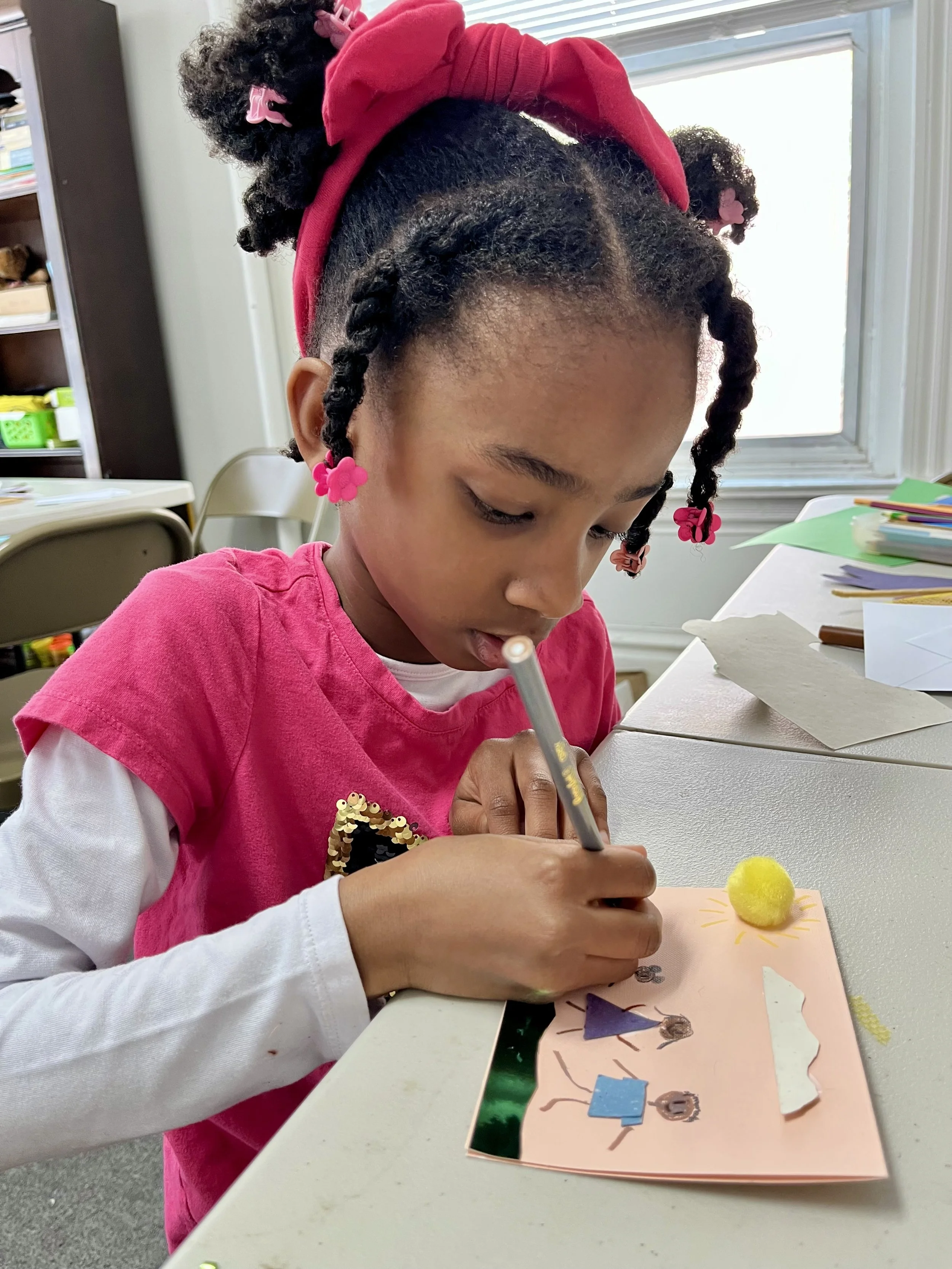 A young girl with braided hair, pink bows, and a red headband is drawing on a pink card with a marker. She is wearing a pink shirt over a white long-sleeve shirt. She is sitting at a table surrounded by art supplies in a classroom.
