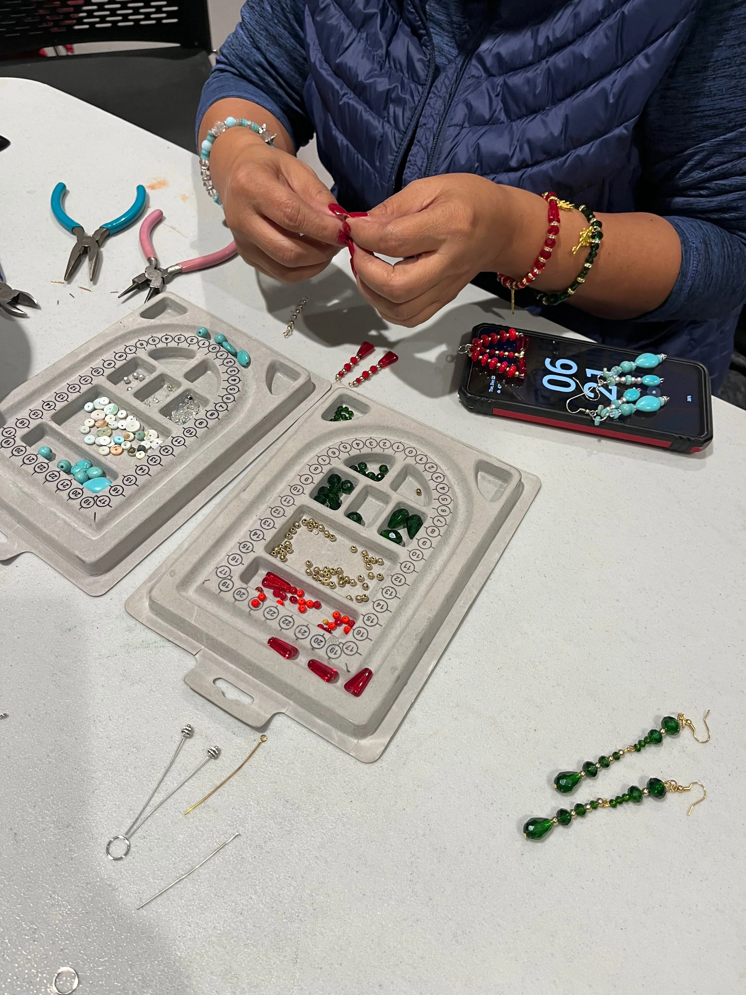 Person making jewelry at a workspace with various beads, pliers, and jewelry tools, wearing multiple colorful beaded bracelets and working with red beads.