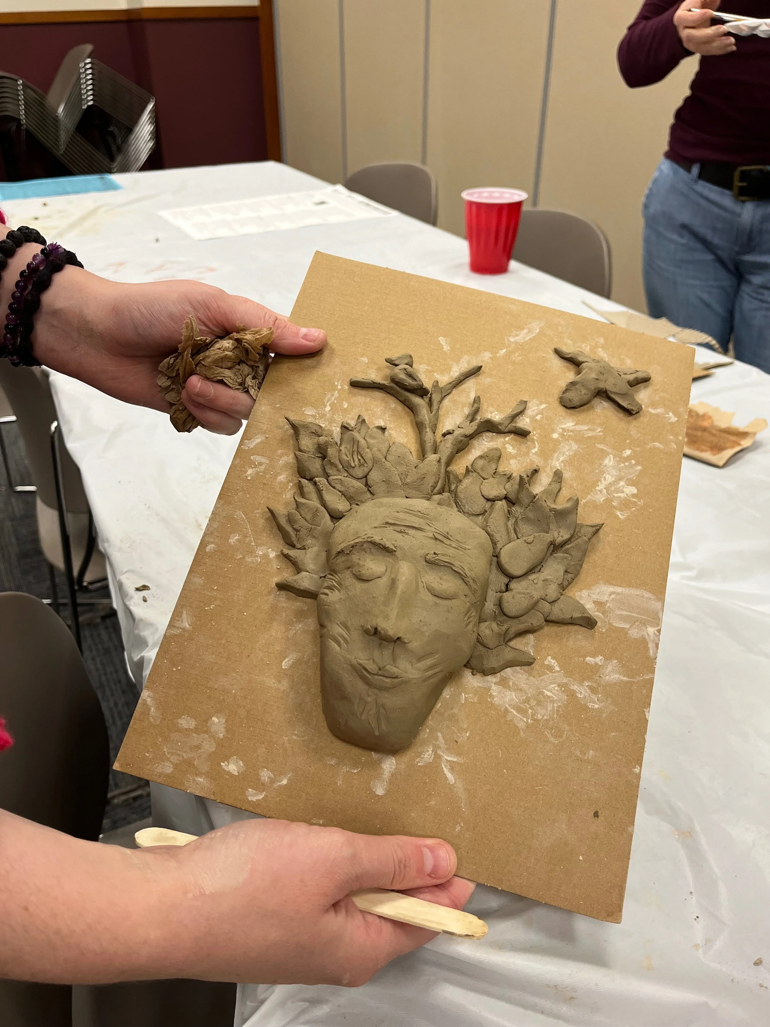 A person holding a clay sculpture of a face with closed eyes, surrounded by leaves and a butterfly, on a piece of cardboard.
