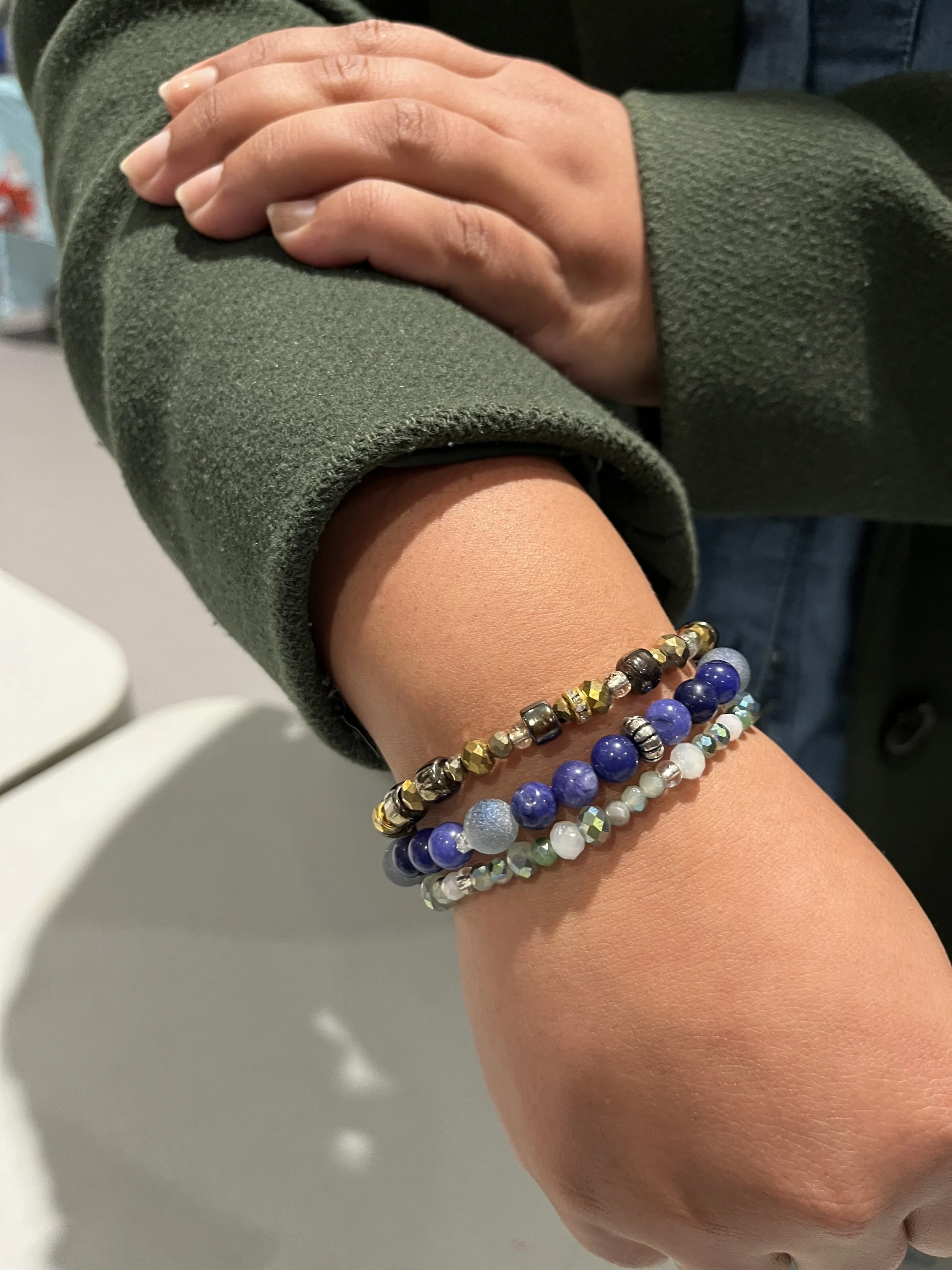 Close-up of a person's wrist with three beaded bracelets, one with blue and white beads, another with metallic and black beads, and a third with clear, white, and iridescent beads, against a background of a person's green sleeve.