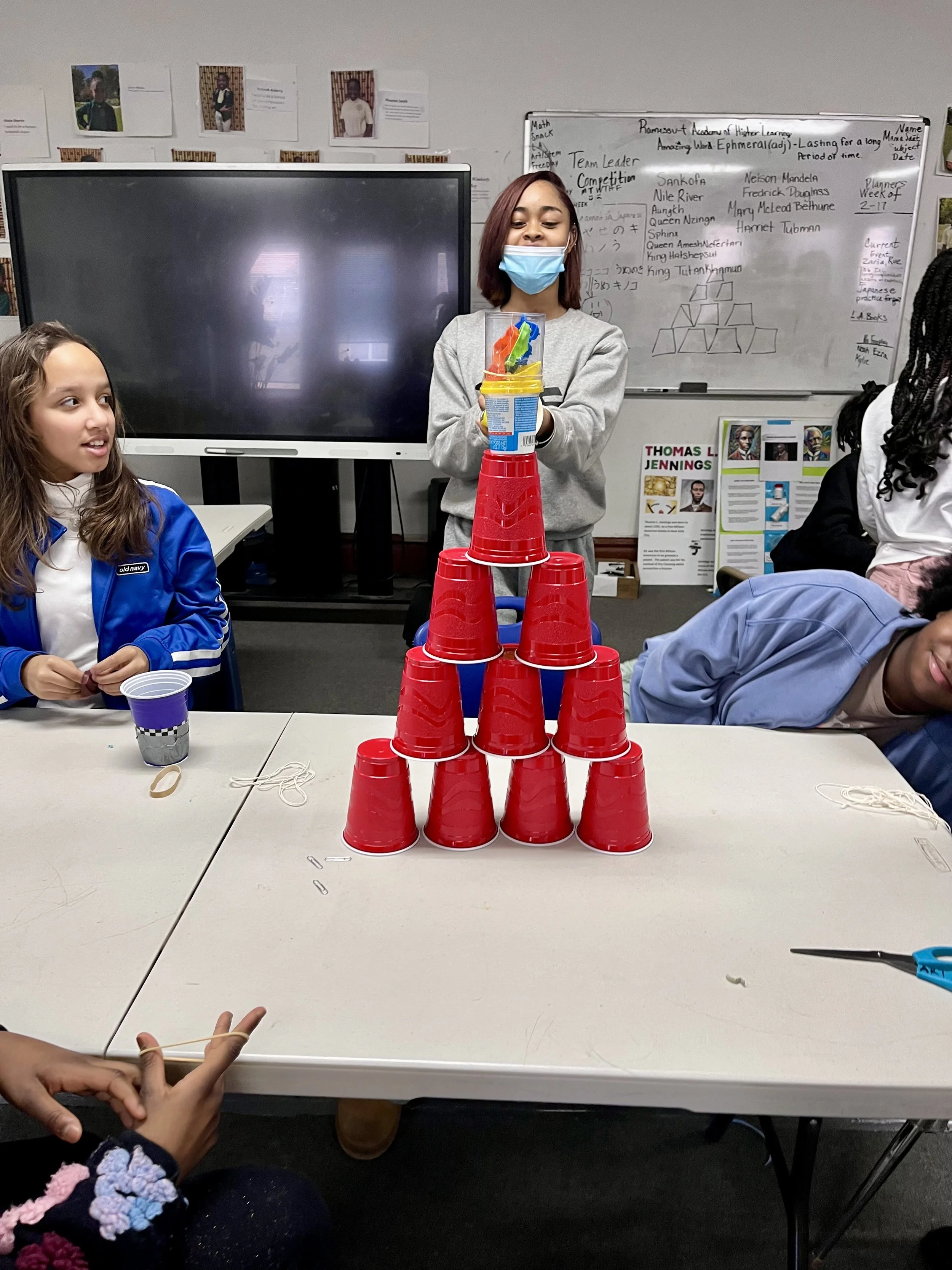 Children participating in a game or activity involving stacking red cups into a pyramid, with one girl wearing a face mask and standing behind the cups, and other children sitting around the table, in a classroom setting.