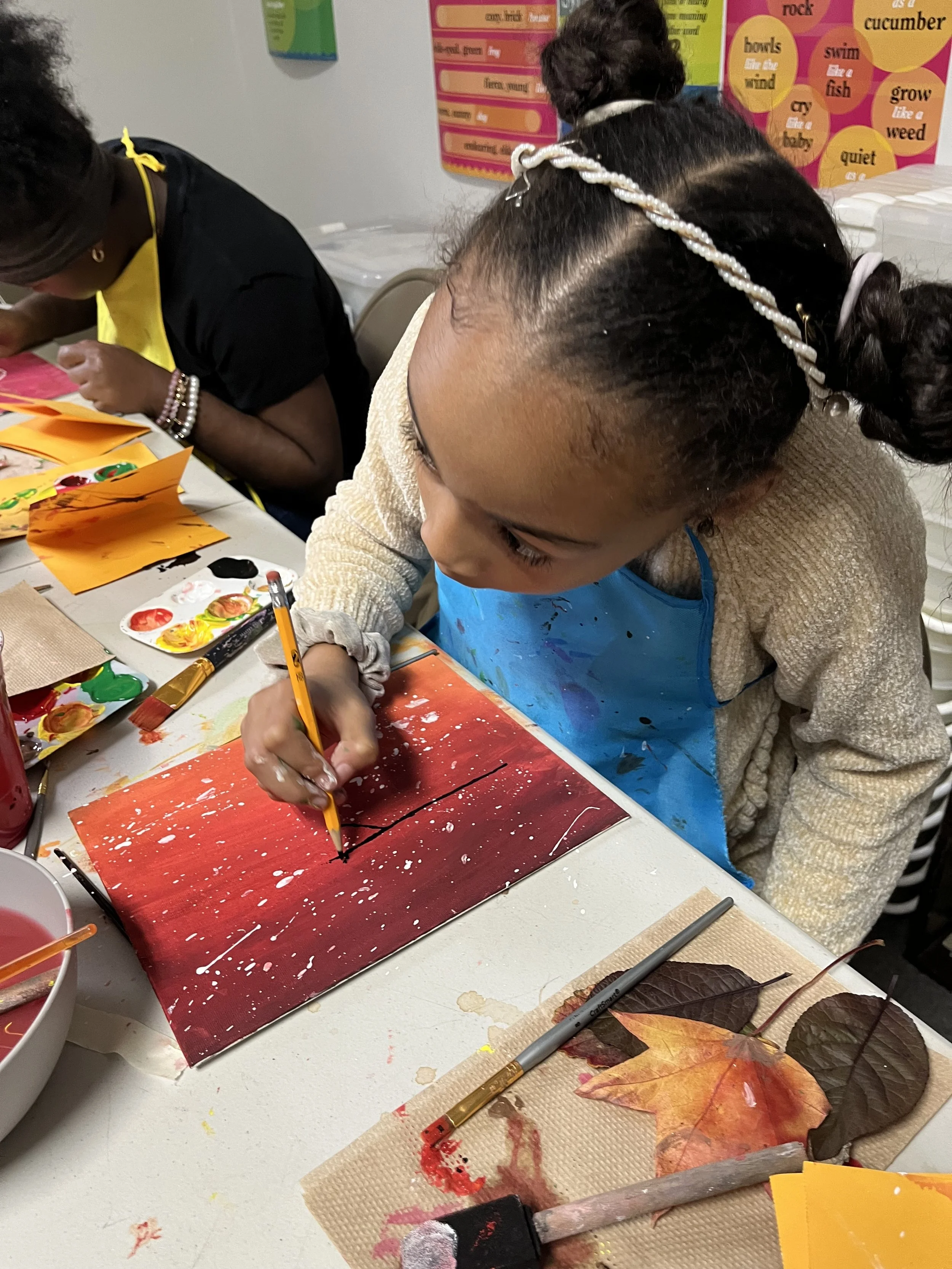 Young girl painting on a canvas with red and white splatters, surrounded by leaves, brushes, and paint supplies on a classroom table.