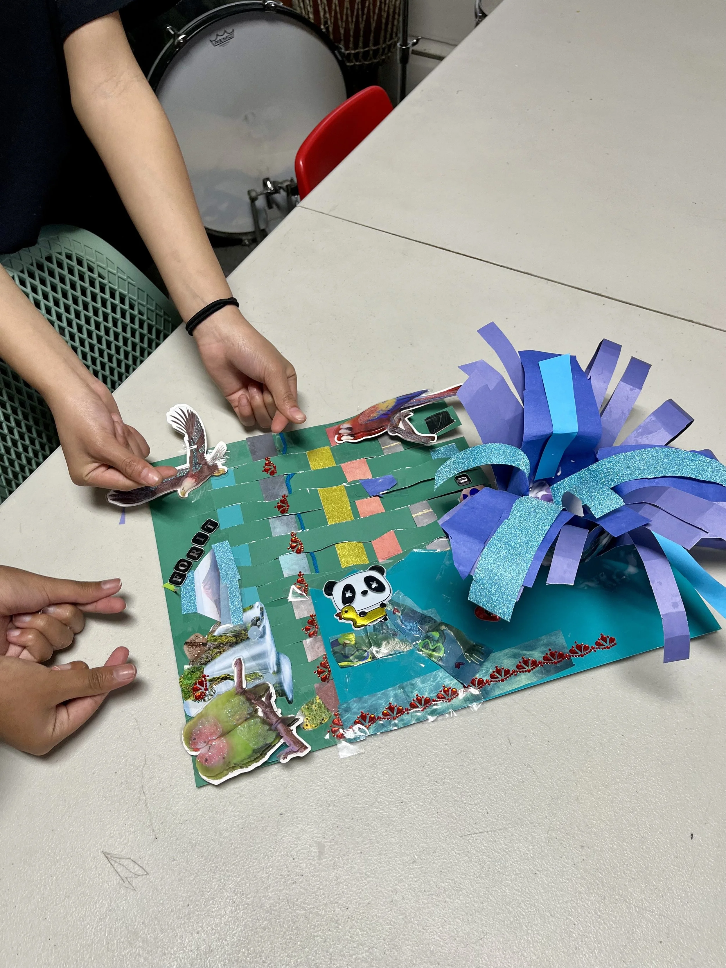 A colorful 3D paper craft project on a table, featuring a woven green paper mat, cutout animal stickers, and purple and blue paper strips. Two people are arranging the craft, with hands visible.