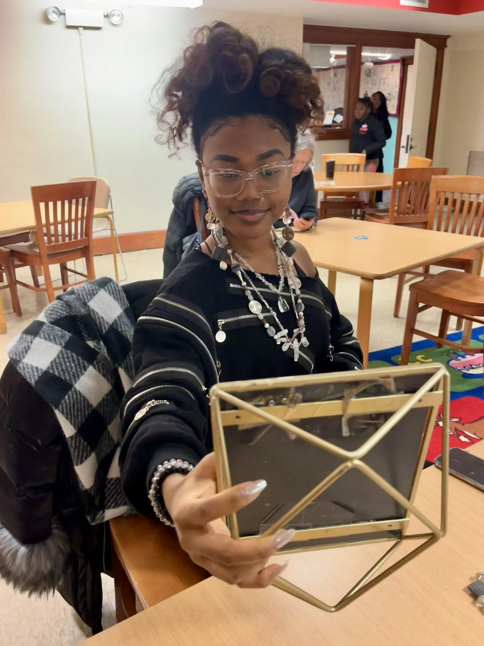 Woman with curly hair styled in buns, wearing clear glasses, layered necklaces, and a black outfit with zipper details, looking at a mirror held in her hand in a room with wooden tables and chairs.
