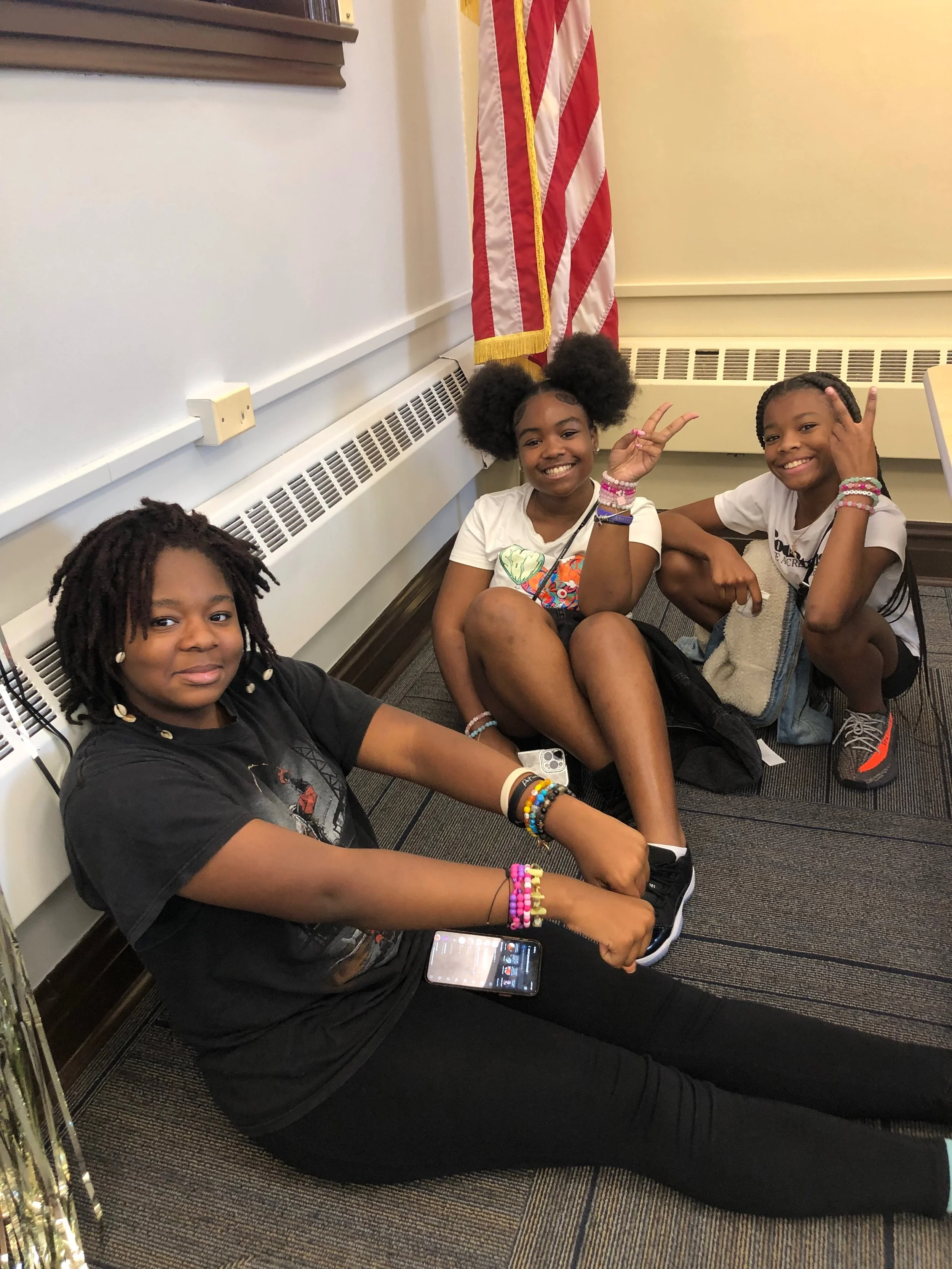 Three young girls sitting and crouching on the floor in a room with a white wall, radiator, and American flag. The girl on the left has dreadlocks, is wearing a black shirt and black pants, and has a smartphone and colorful bracelets. The girl in the