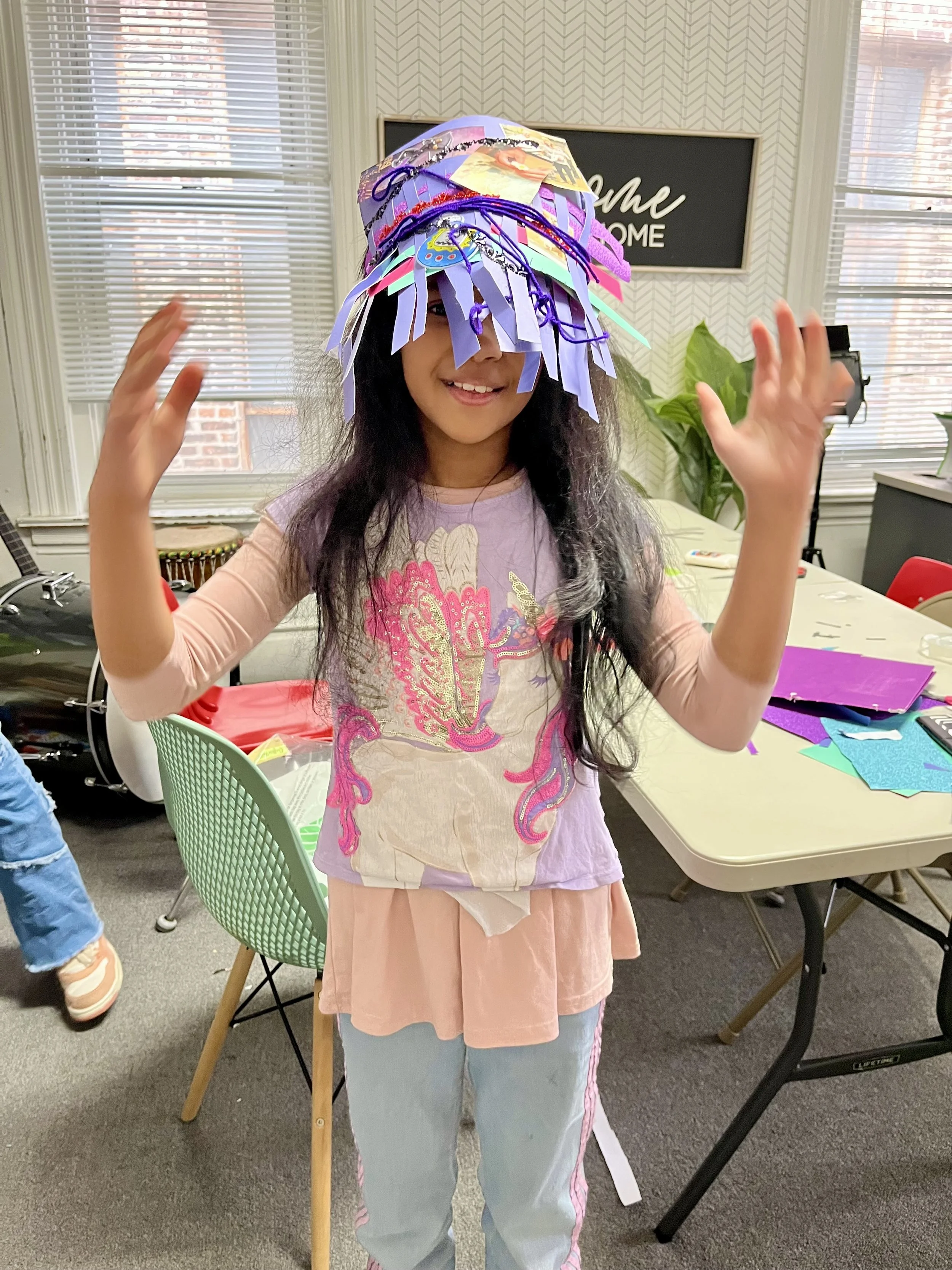 A young girl with long dark hair wearing a colorful paper craft hat, smiling and raising her hands in a room with large windows, a table with craft supplies, and a "Home" sign on the wall.