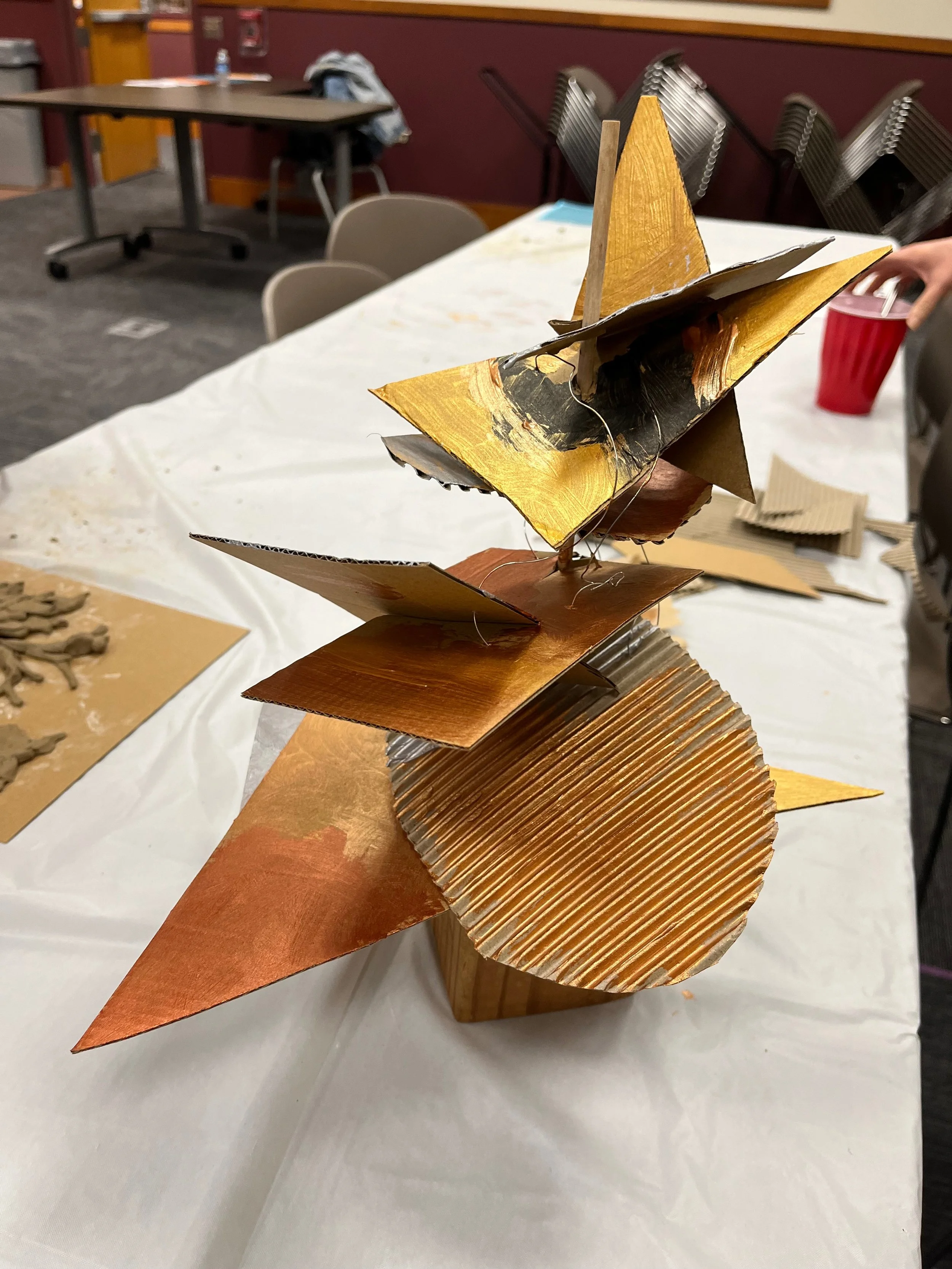 Metal sculpture with geometric shapes in gold, bronze, and silver, arranged on a wooden base, placed on a white tablecloth in an indoor setting.