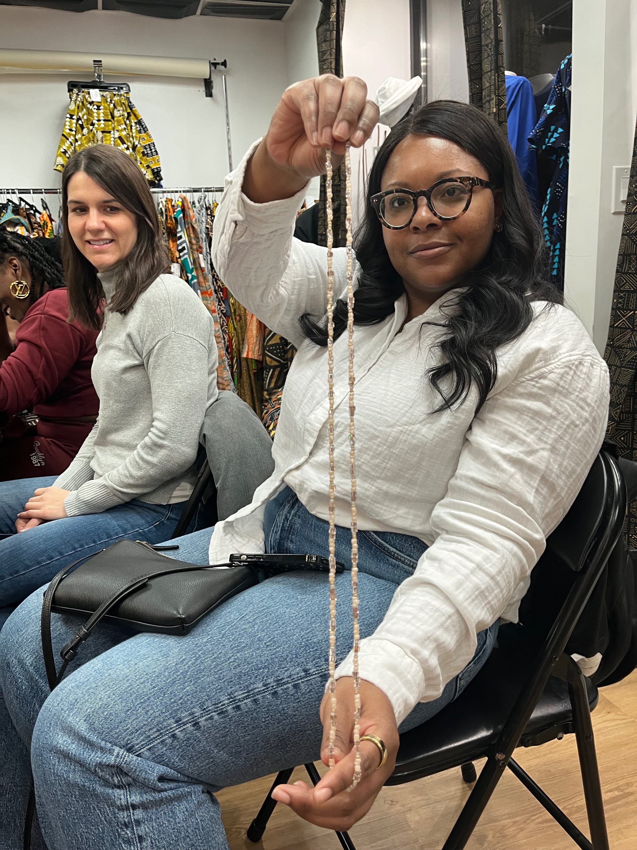 A woman with black hair, glasses, and a white shirt holding a necklace in a room with other people and colorful clothing racks.