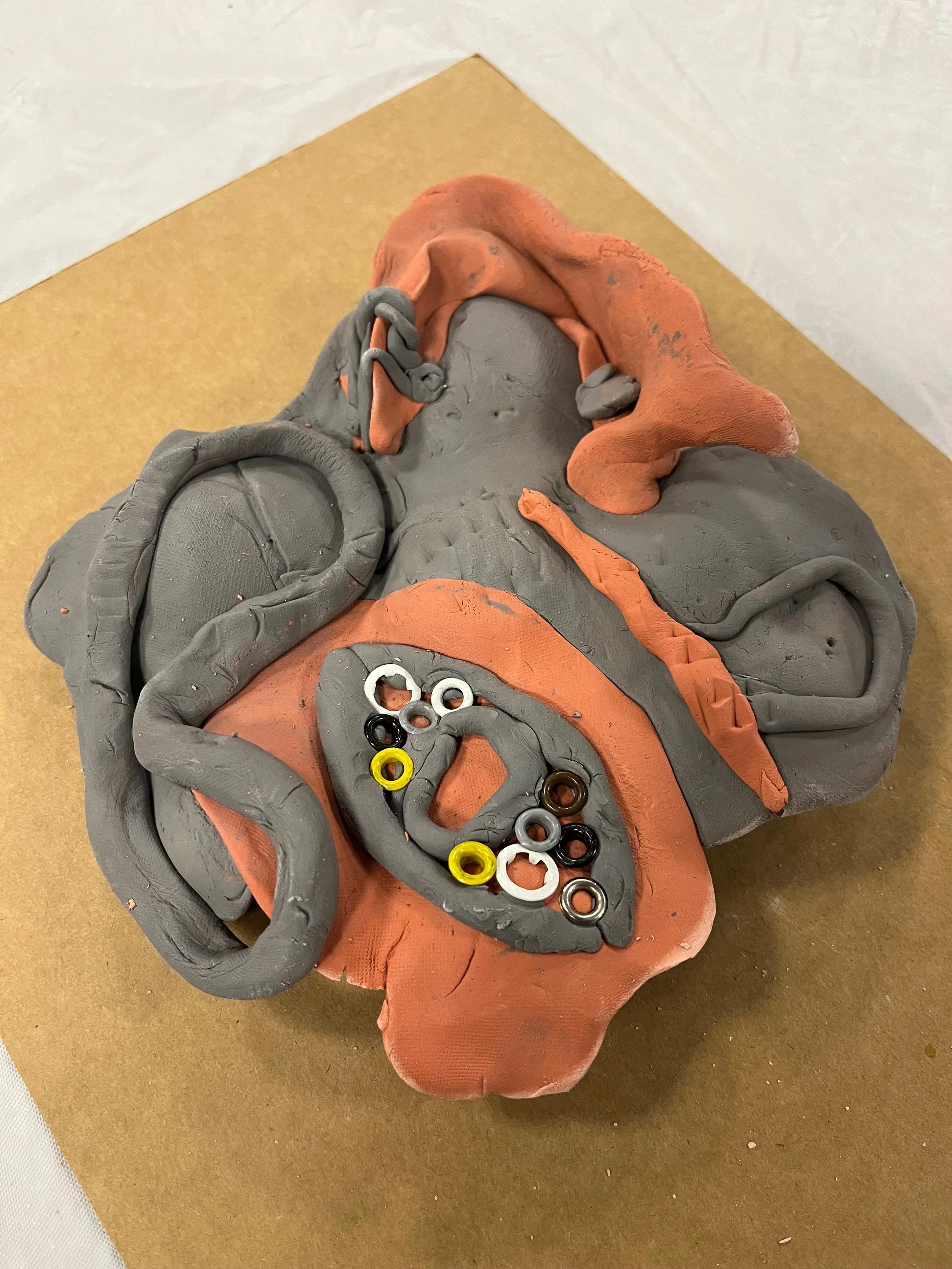 A sculpture of a human head made from gray and orange clay with various small rings embedded in an open mouth.