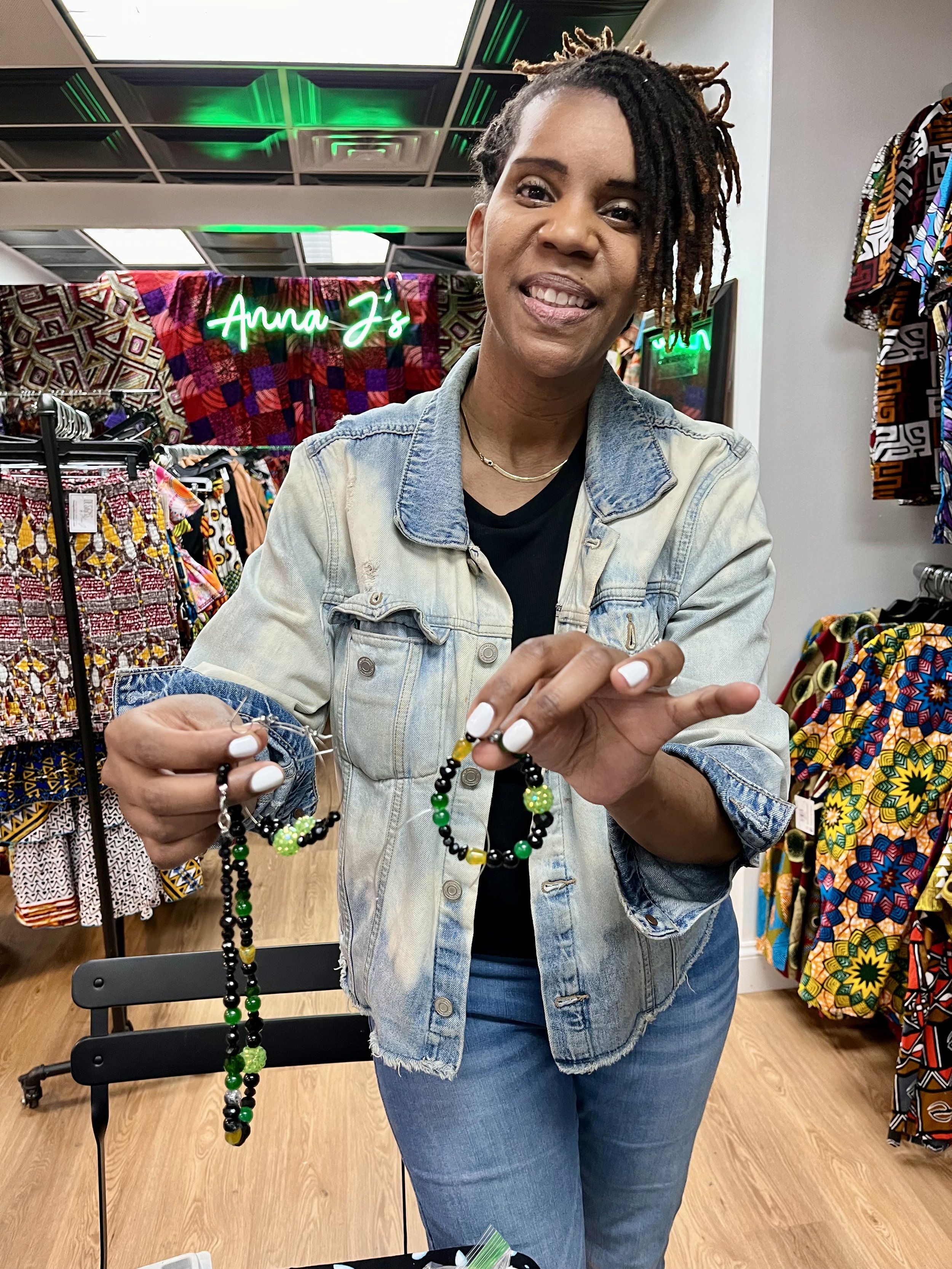 A woman with short dreadlocks wearing a denim jacket standing in a store, holding colorful beaded jewelry, with clothing racks and a neon sign reading 'Anna J's' in the background.