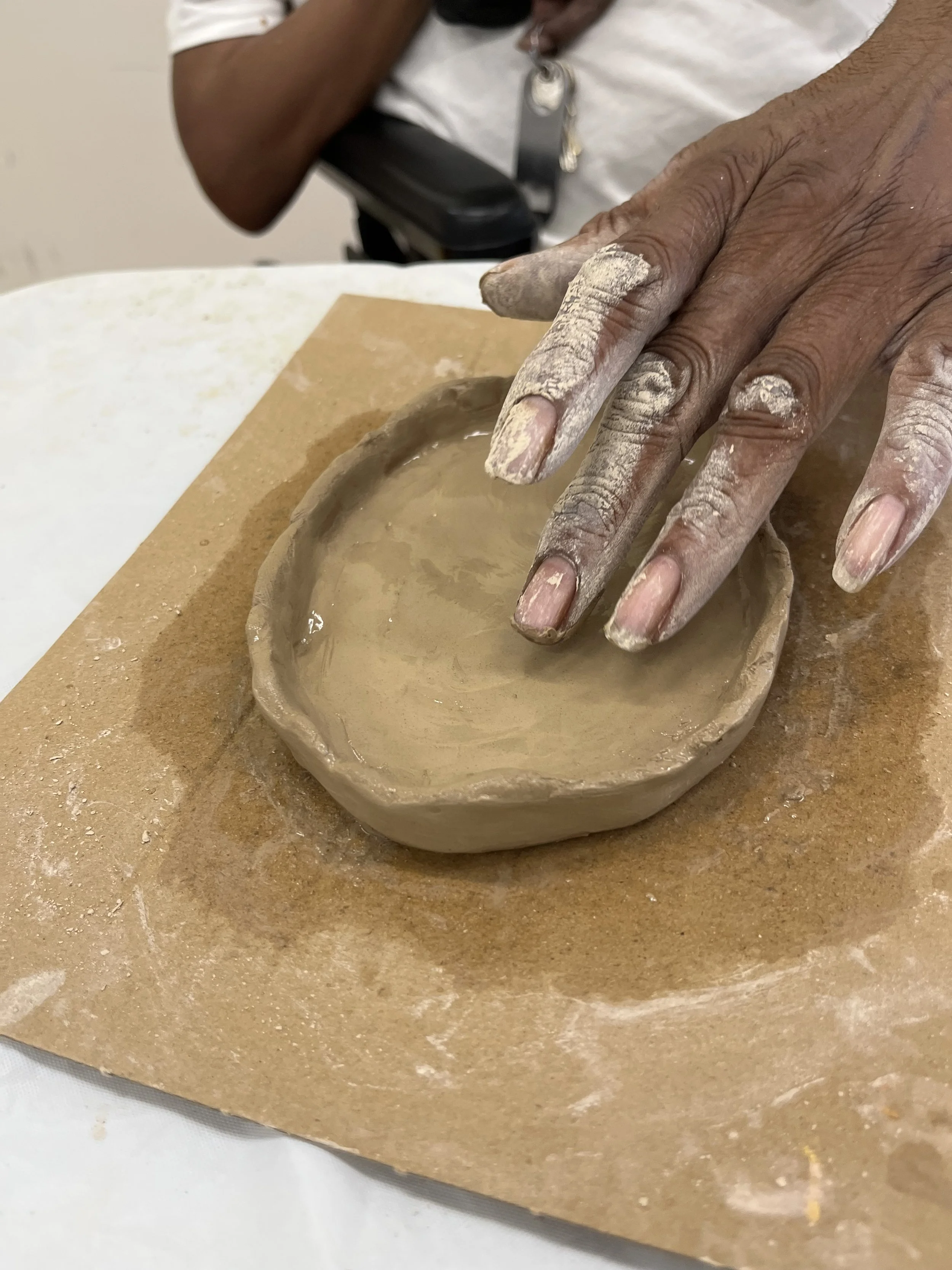 A person with clay-covered hands shaping a small clay bowl on a piece of cardboard.