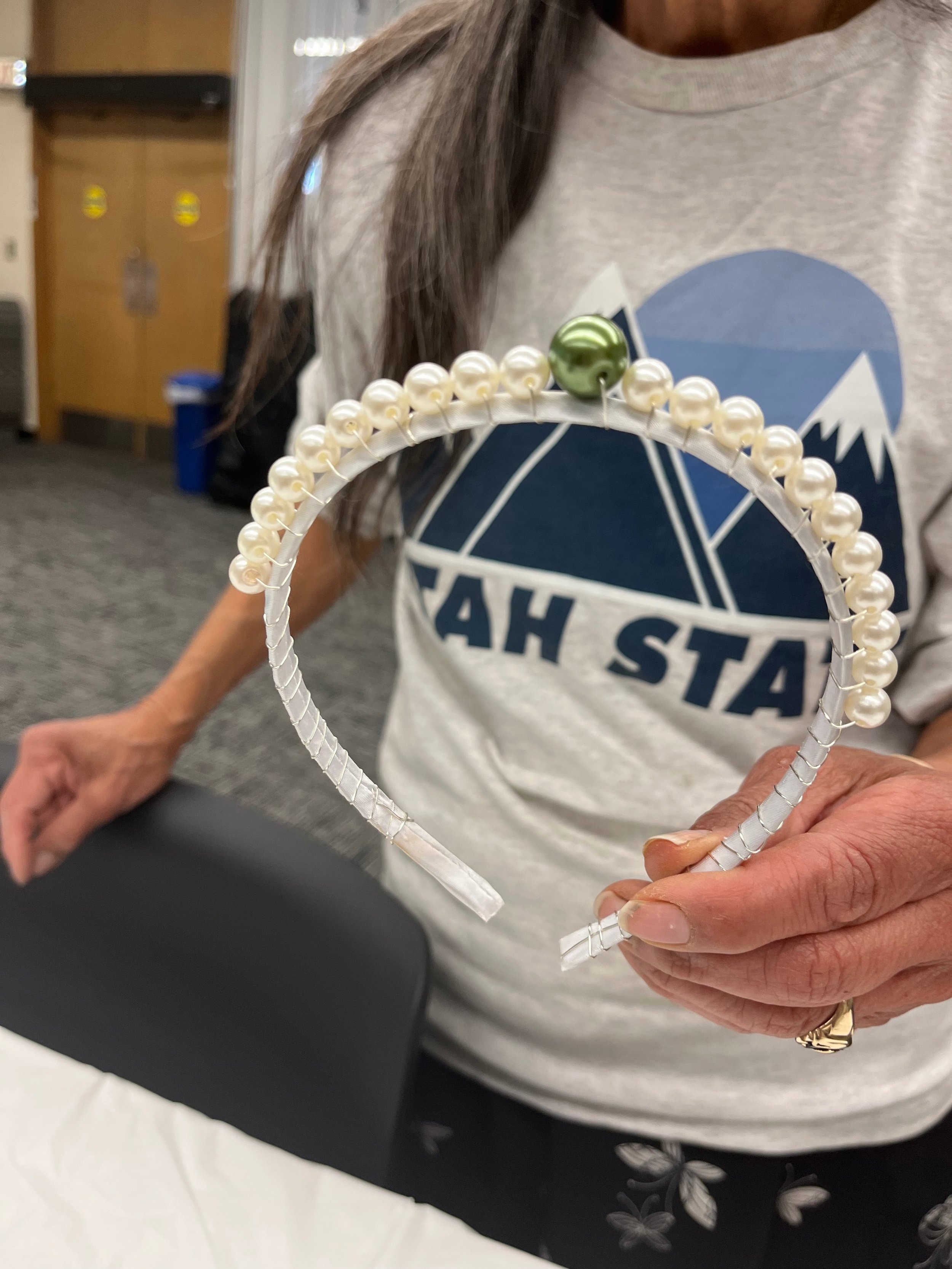 Person holding a pearl-studded white headband with a green decorative bead, wearing a gray shirt with a mountain and logo design.