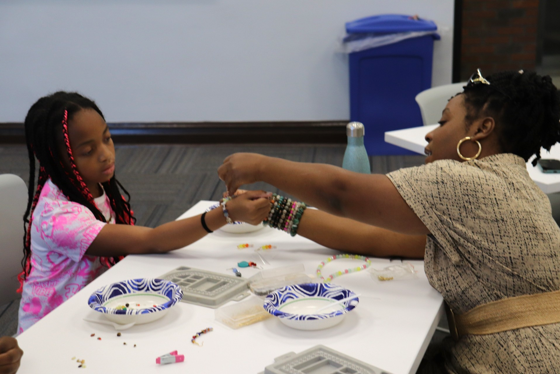 Woman assisting a young girl with beading jewelry at a table with beads, bowls, and craft supplies.