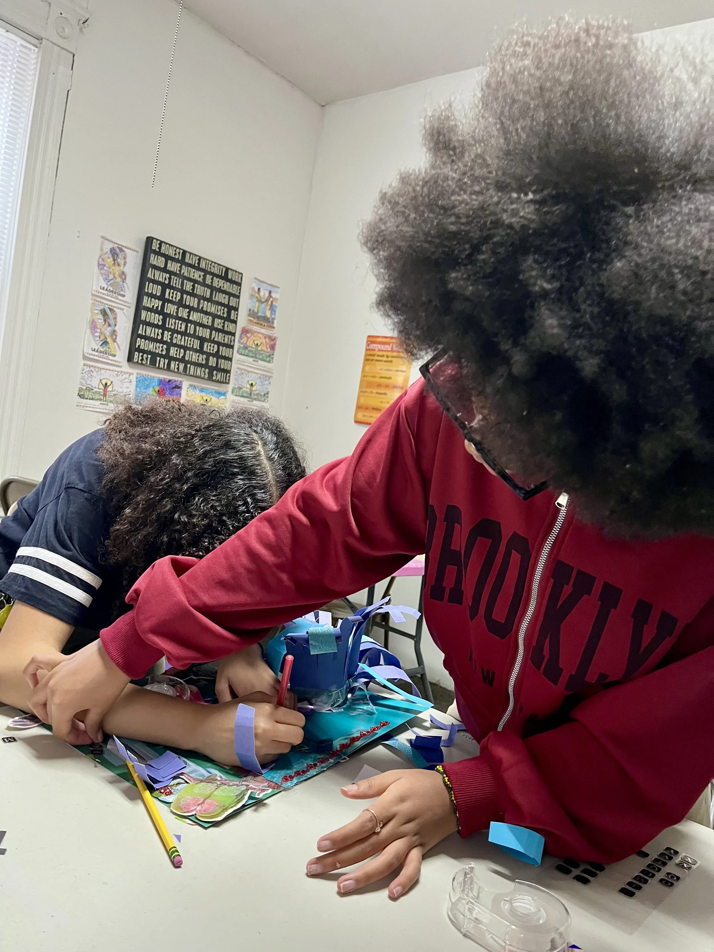 Two girls are working on a arts and crafts project at a table, with one girl writing and the other girl assisting. The table has craft supplies and decorations.