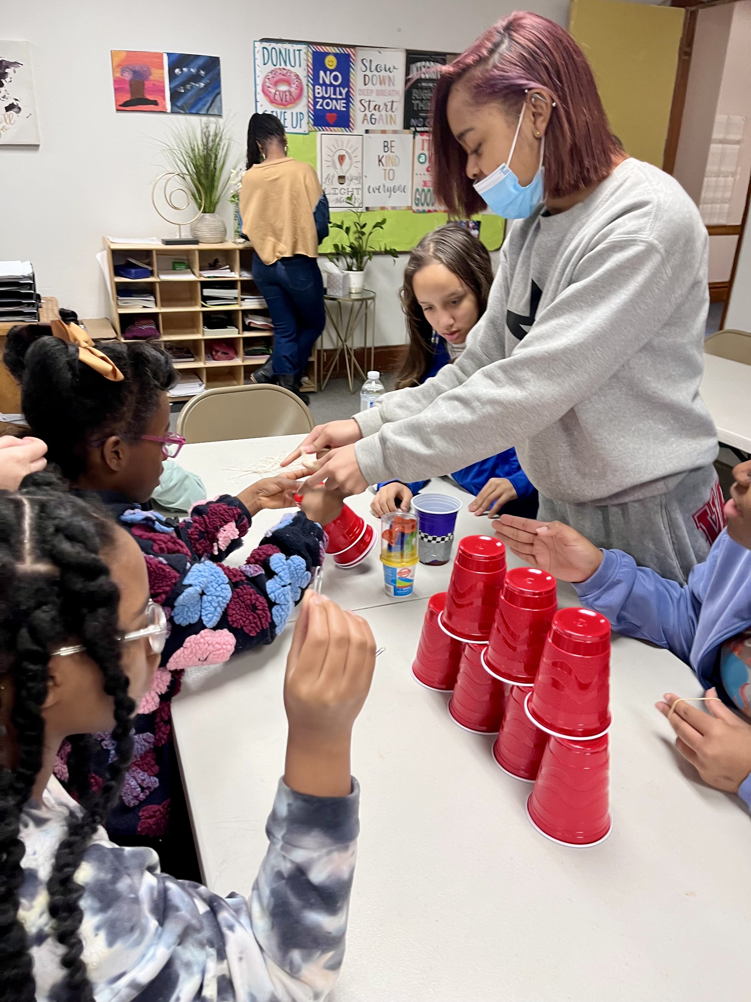 An adult wearing a face mask and gray sweatshirt is helping children with a stacking game involving red plastic cups on a table. Several children are participating, some with glasses and colorful clothing, in a classroom with posters and plants in the background.