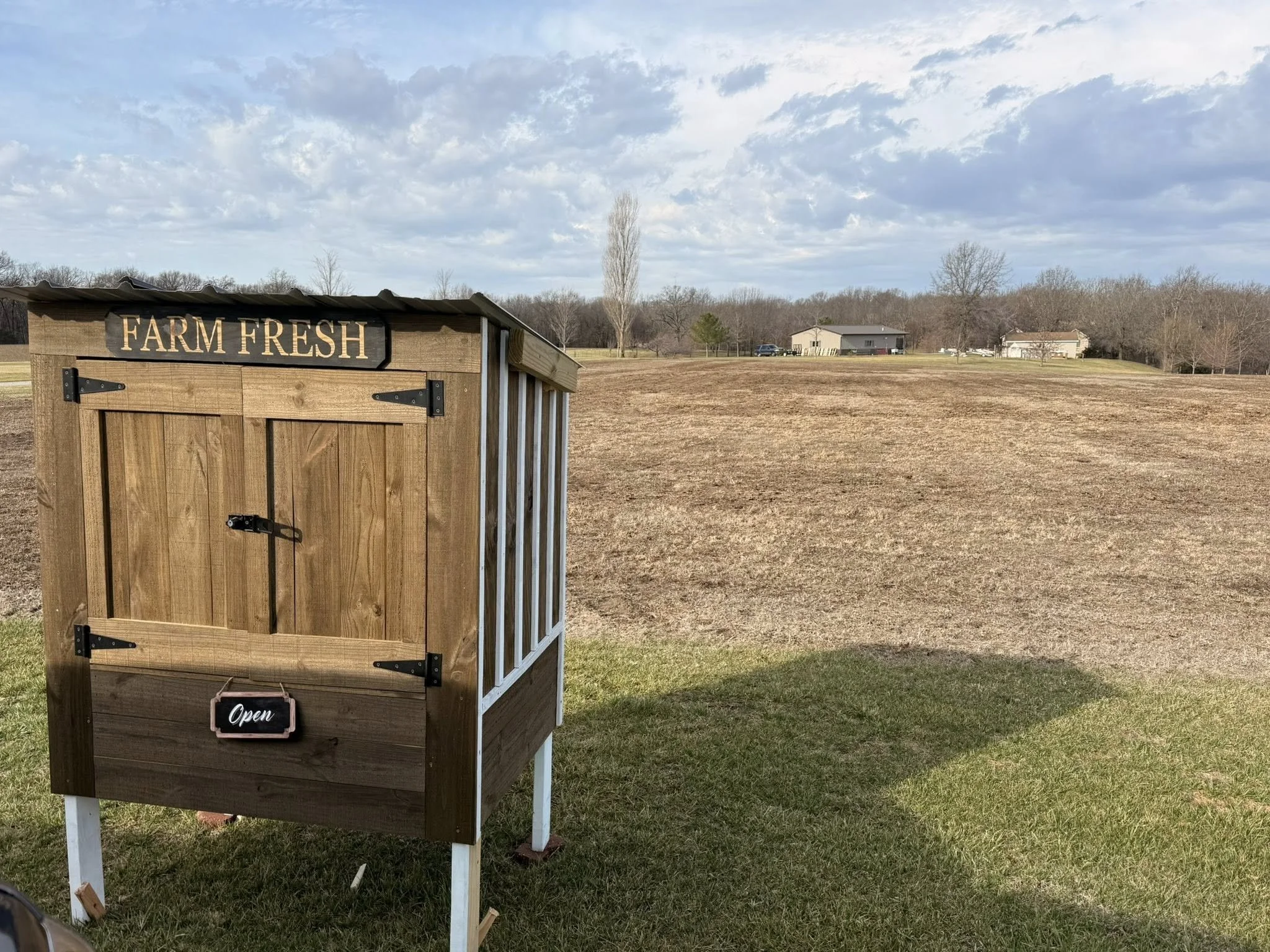 Wooden farm stand with a sign that reads 'FARM FRESH' and a small 'Open' sign hanging from the door, situated on grass with a field and houses in the background under a blue sky with clouds.