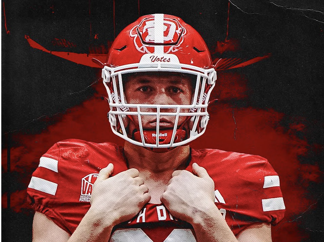 Young male football player wearing maroon and yellow jersey with the number 32, holding a football helmet over his shoulders, with a dark background and orange lighting.