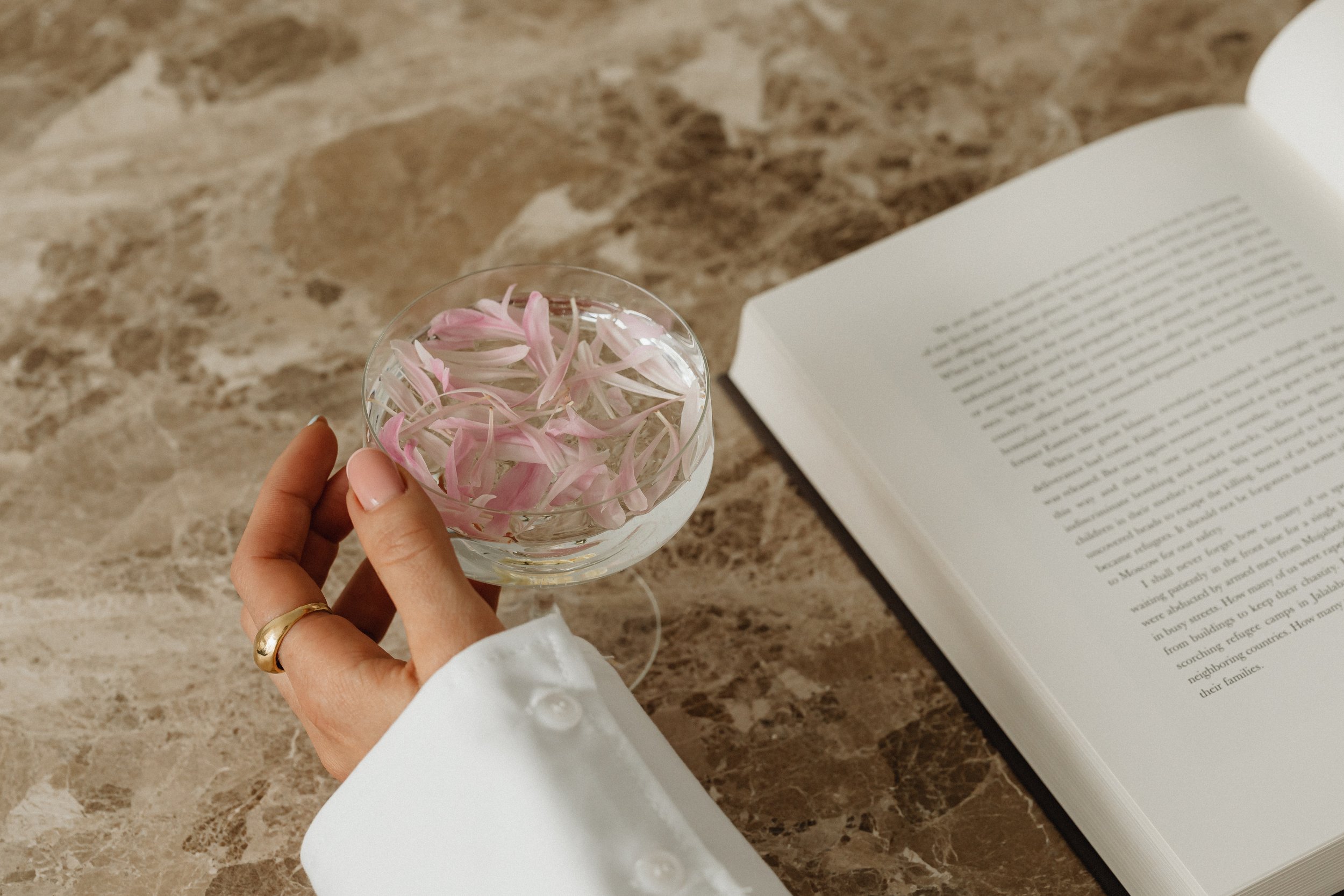 A person's hand with a gold ring holding a glass bowl of pink and white flower petals over a marble surface next to an open book.