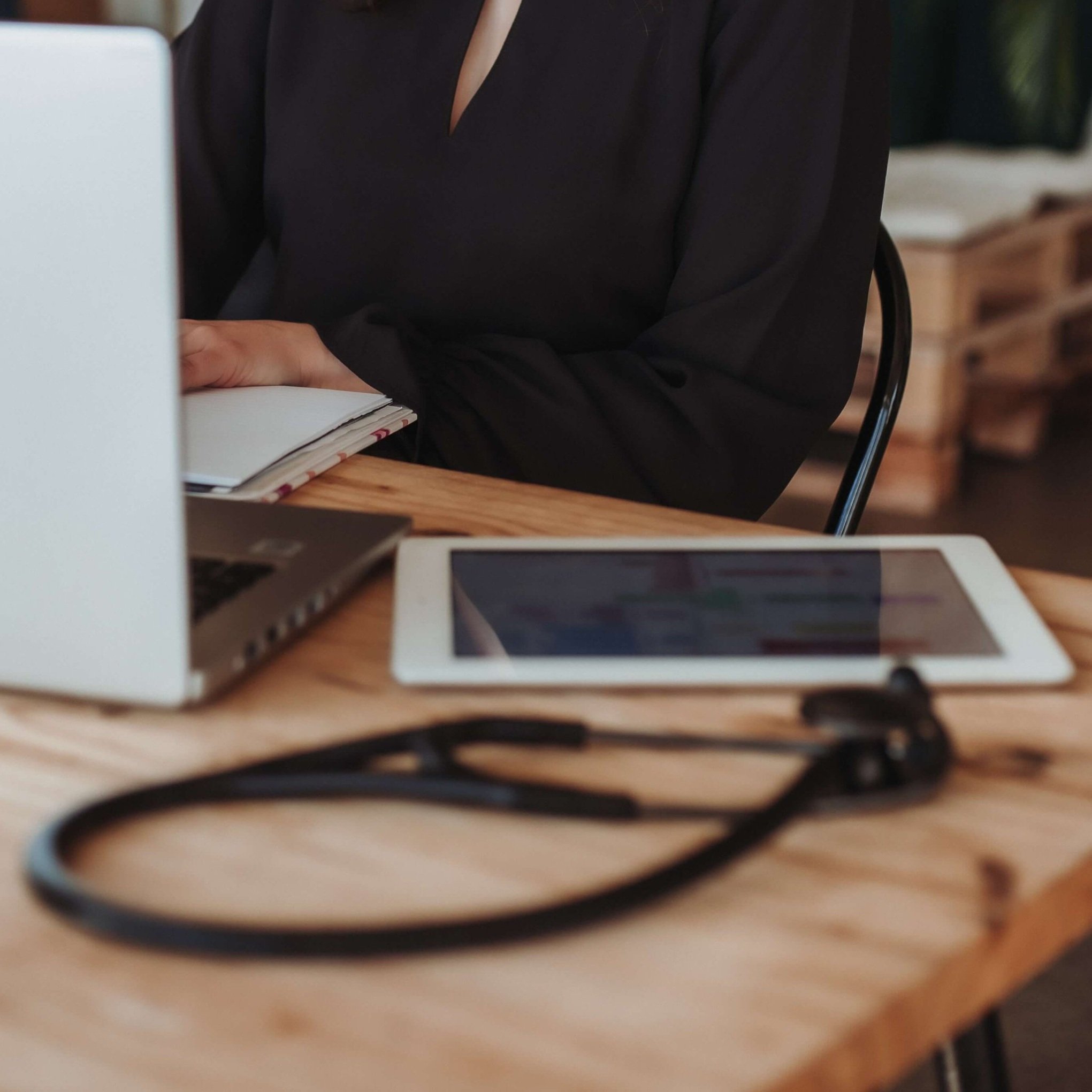 A person sitting at a desk with a laptop, tablet, notebook, and stethoscope.