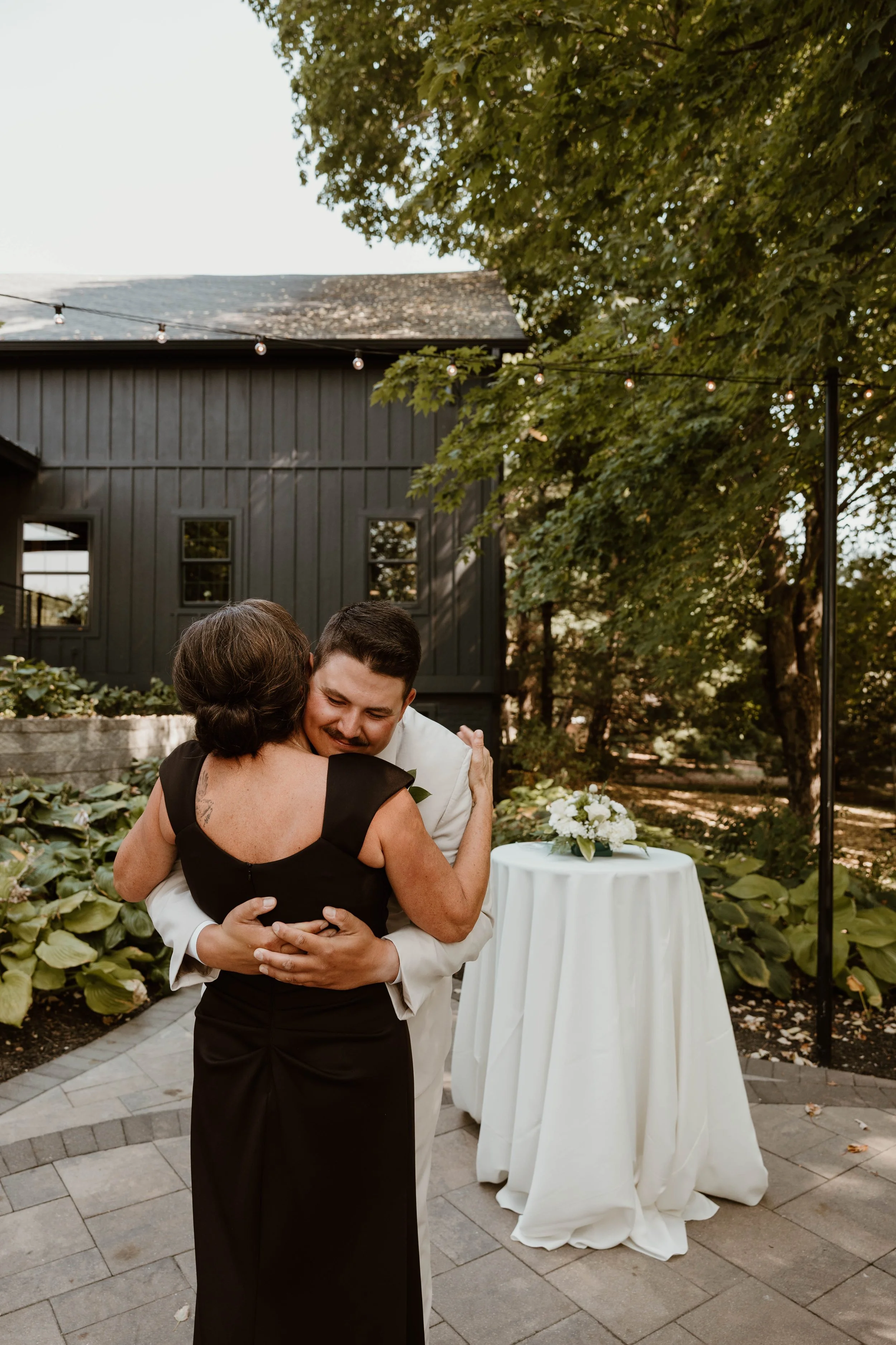 A man and a woman embrace and share a hug outdoors, near a table with a white cloth and floral arrangement, surrounded by trees and string lights.