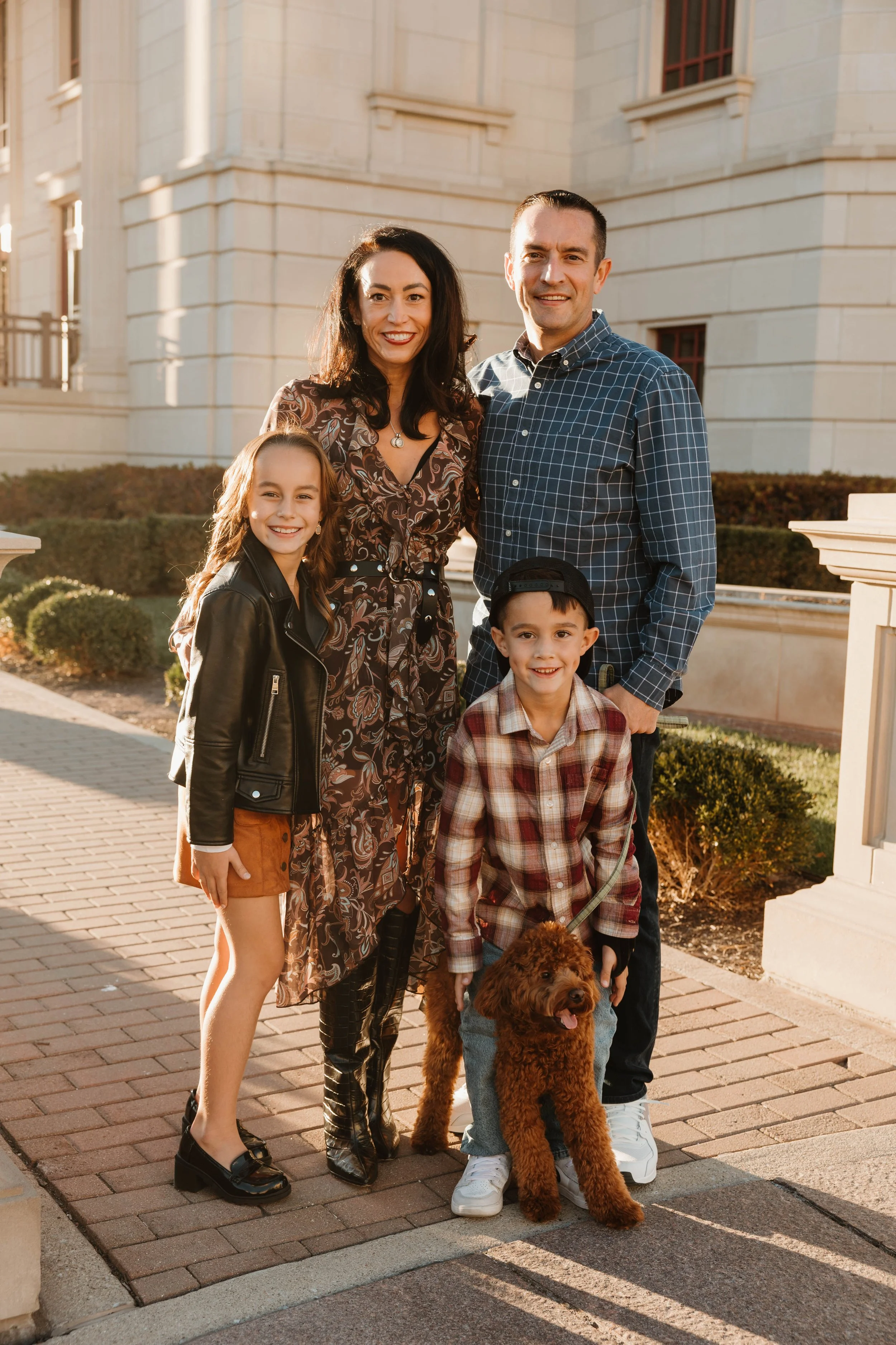 A family of four with a dog outside a building, smiling at the camera in the late afternoon.